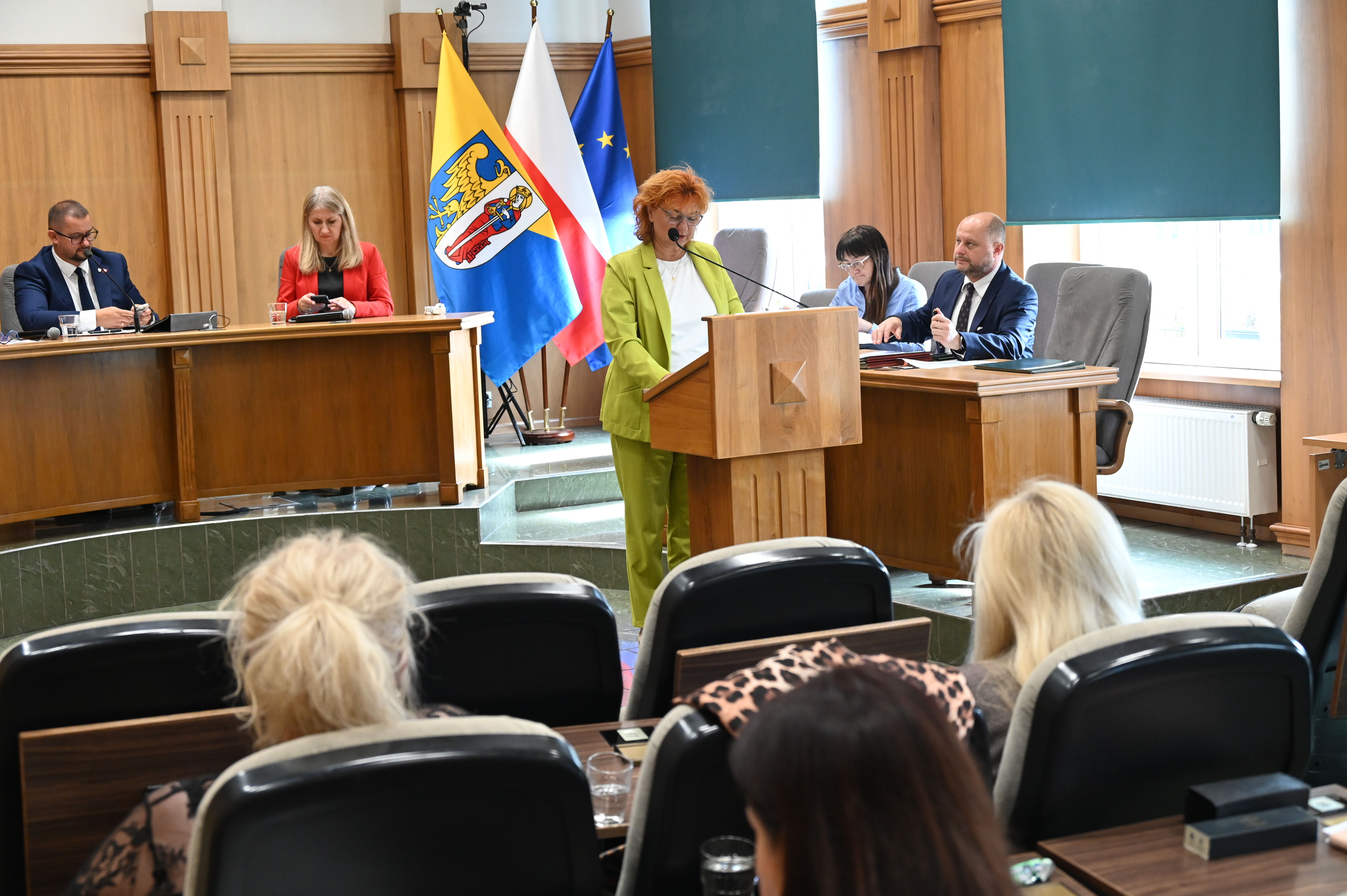 Kobieta przy mównicy, ludzie siedzą na krzesłach.