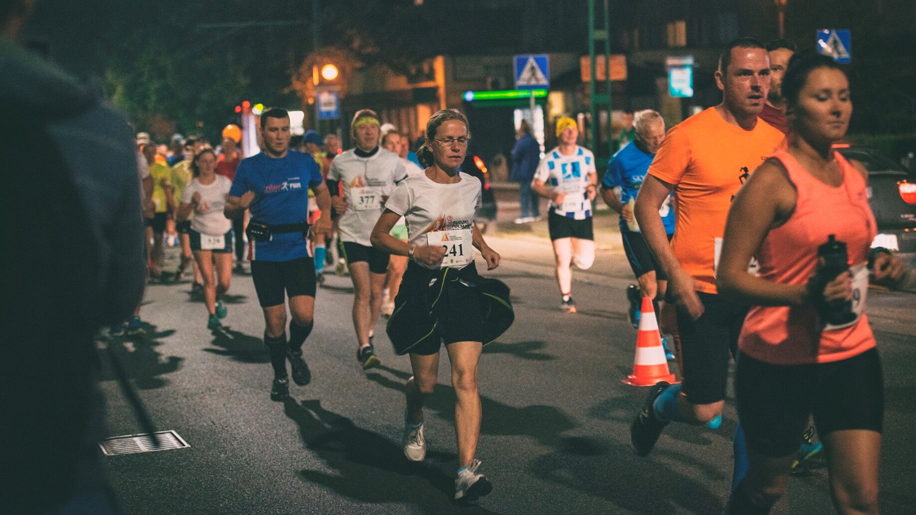 Biegacze z numerami startowymi na ulicy, w tle znaki drogowe, budynki i świecąca latarnia.