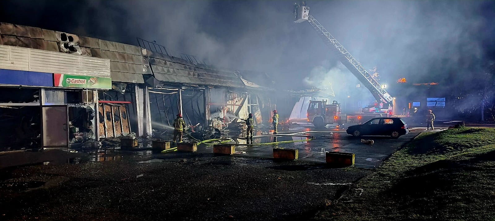Strażacy dogaszający wypalony pawilon handlowy, w tle wóz straży pożarnej z wysięgnikiem.