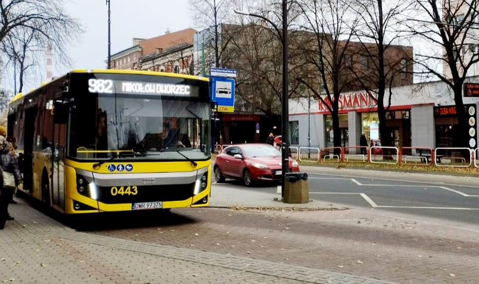 Żółty autobus linii 982 stoi na przystanku. Obok jedzie czerwone auto osobowe. W tle drzewa, sklepy.