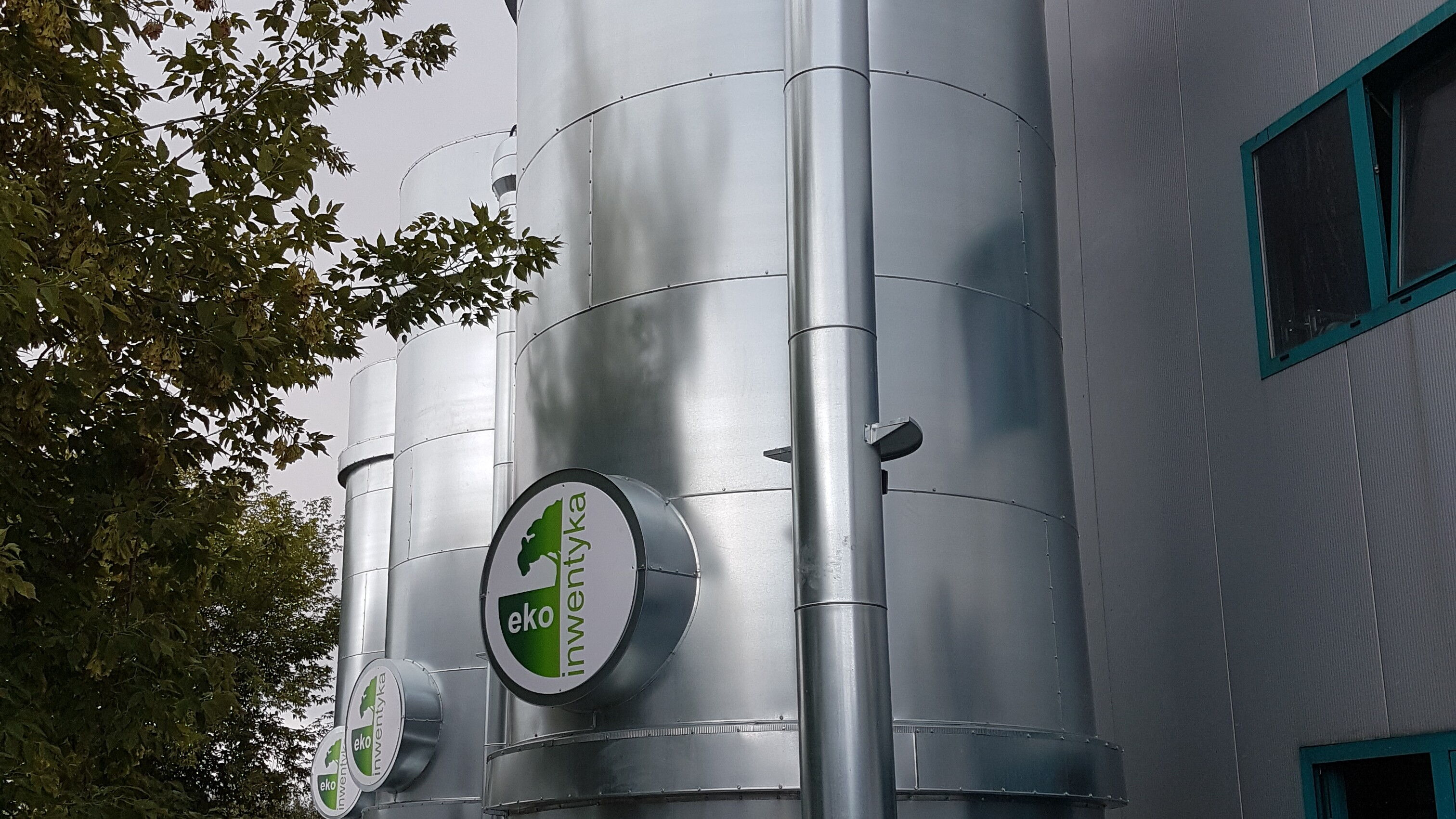 Drzewa, bioreaktory z logo Ekoinwentyka, ściana budynku.