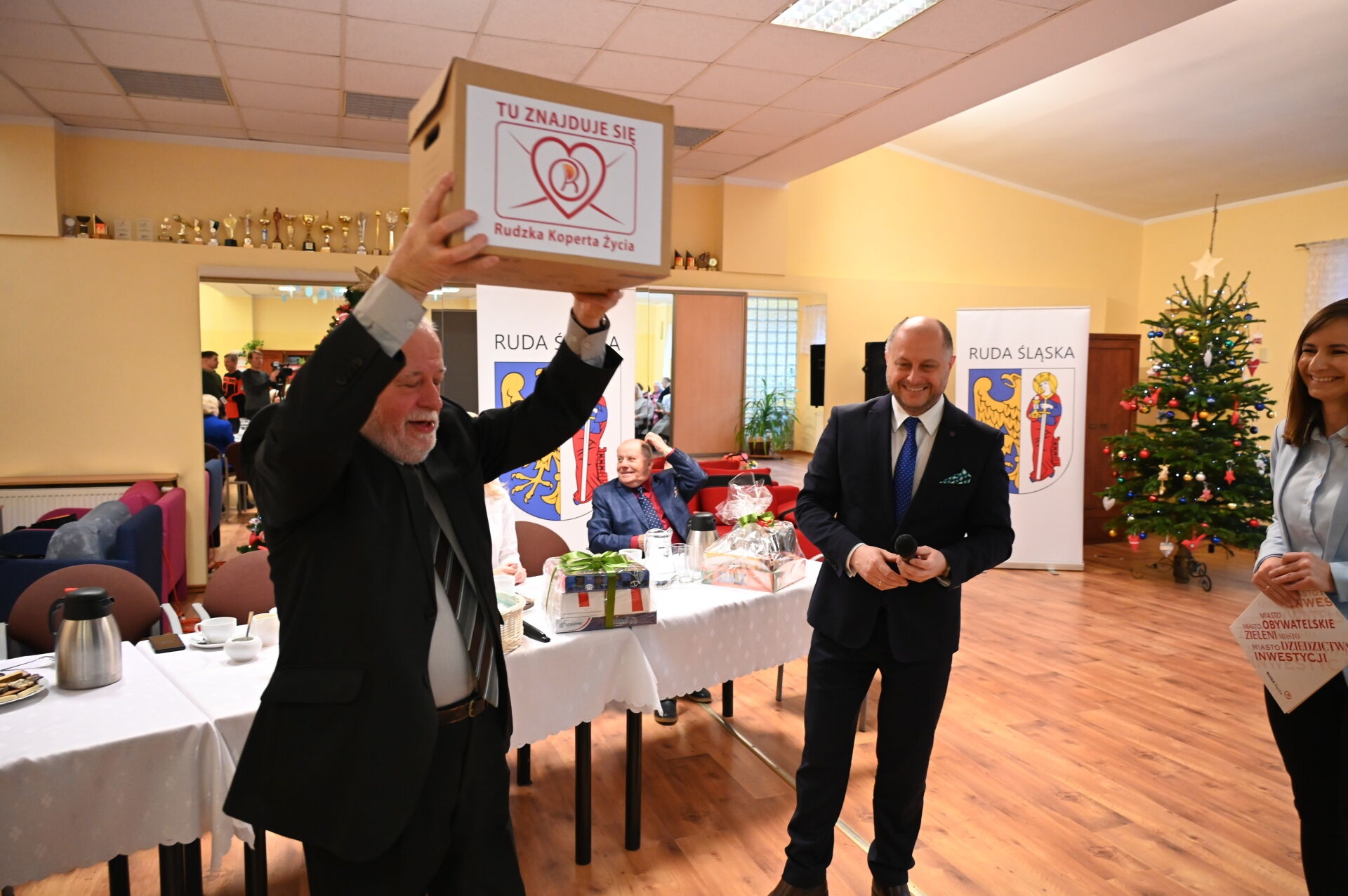 Dwóch mężczyzn i kobieta. Jeden ma podniesione ręce do góry i trzyma w nich karton.