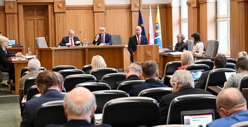 Sala sesyjna. Grupa osób siedzących na fotelach, w tle mężczyzna przy mównicy i kilka osób za stołem prezydialnym. Z tyłu flagi Unii Eurospejskiej, Polski, i Rudy Śląskiej.