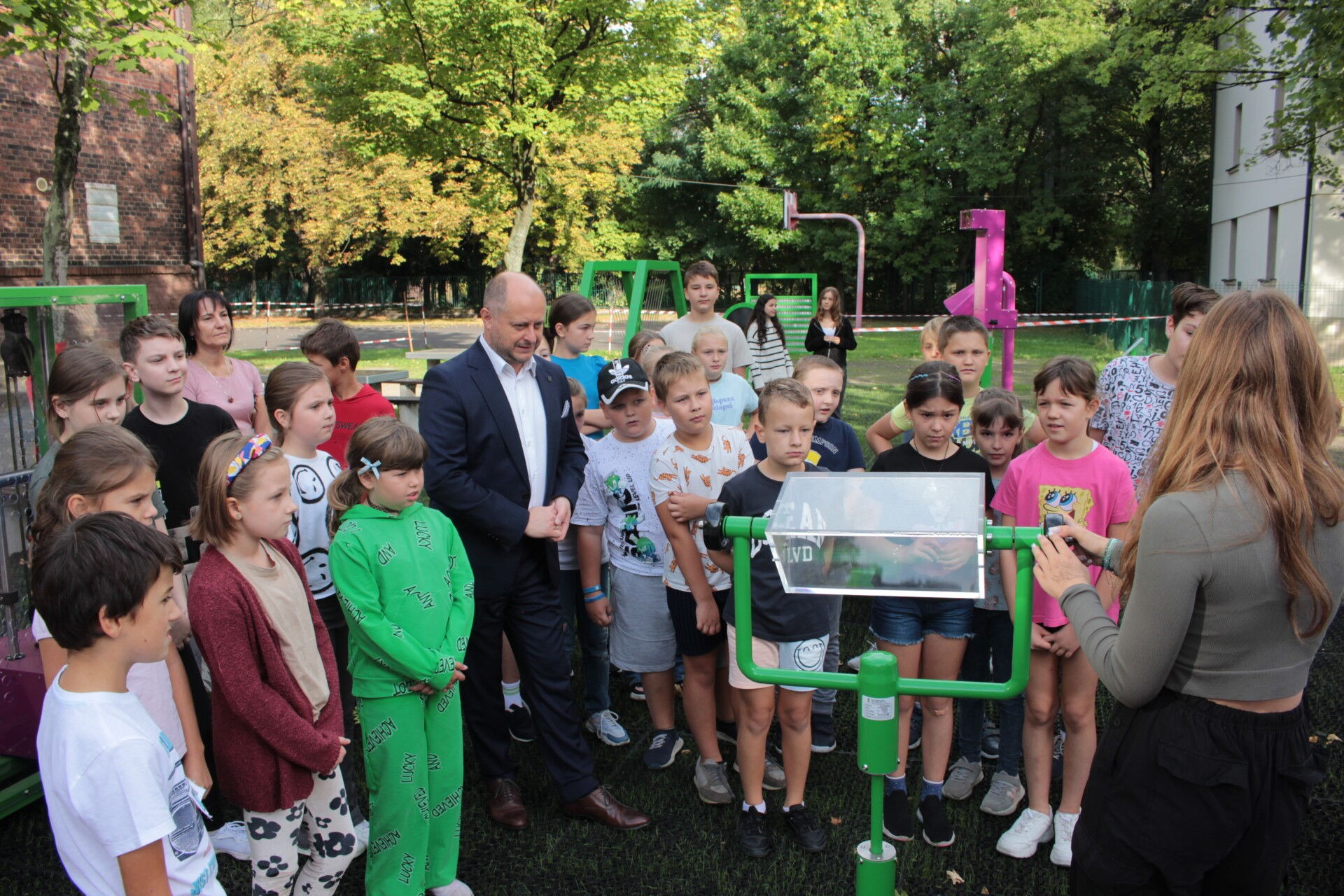 Mężczyzna w garniturze wśród grupy dzieci przygląda się prezentowanemu przez uczennicę pryzmatowi. W tle inne urządzenia, budynki i zieleń.