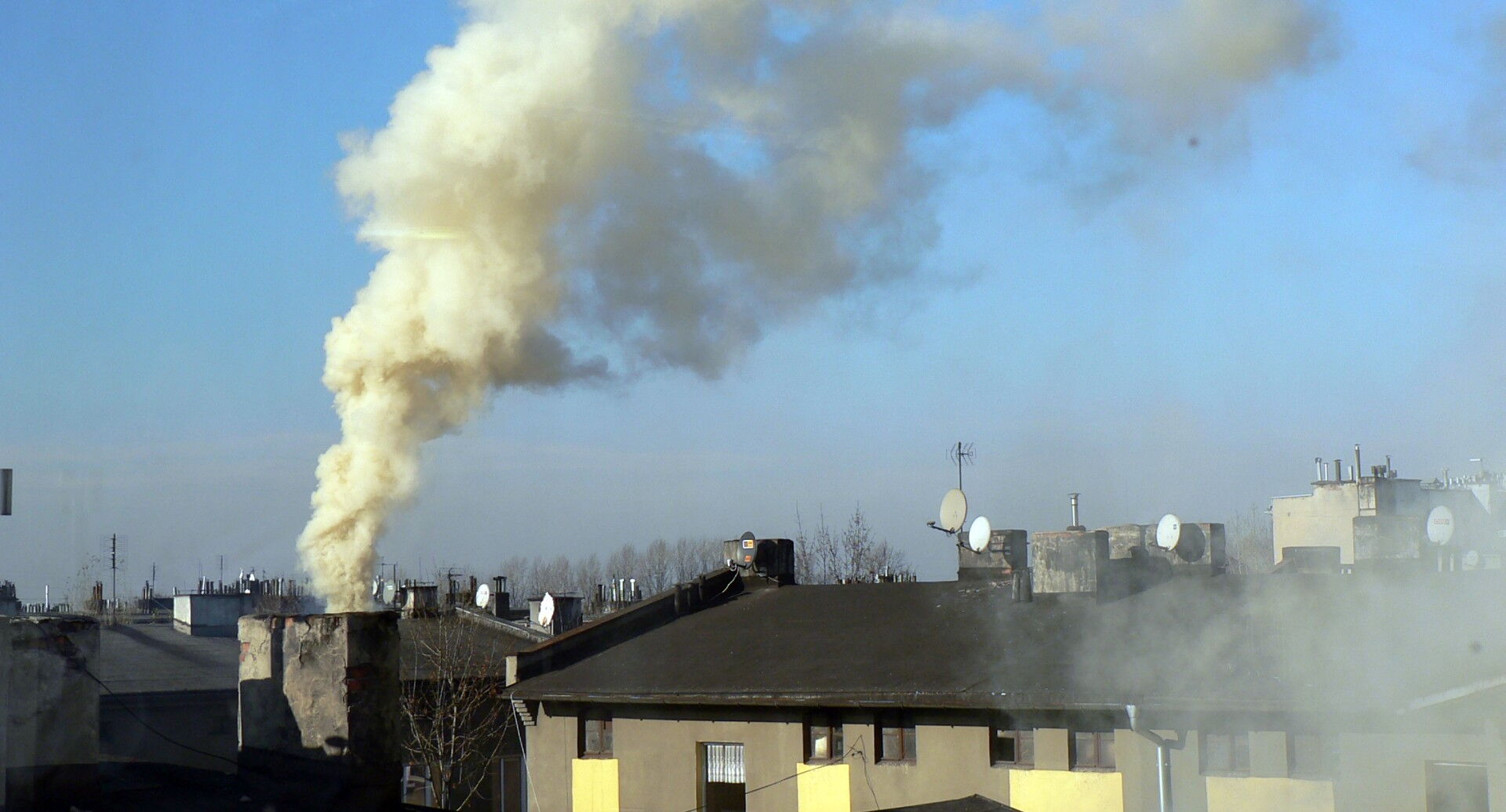 Dymiący komin, w tle dachy budynków i niebo.