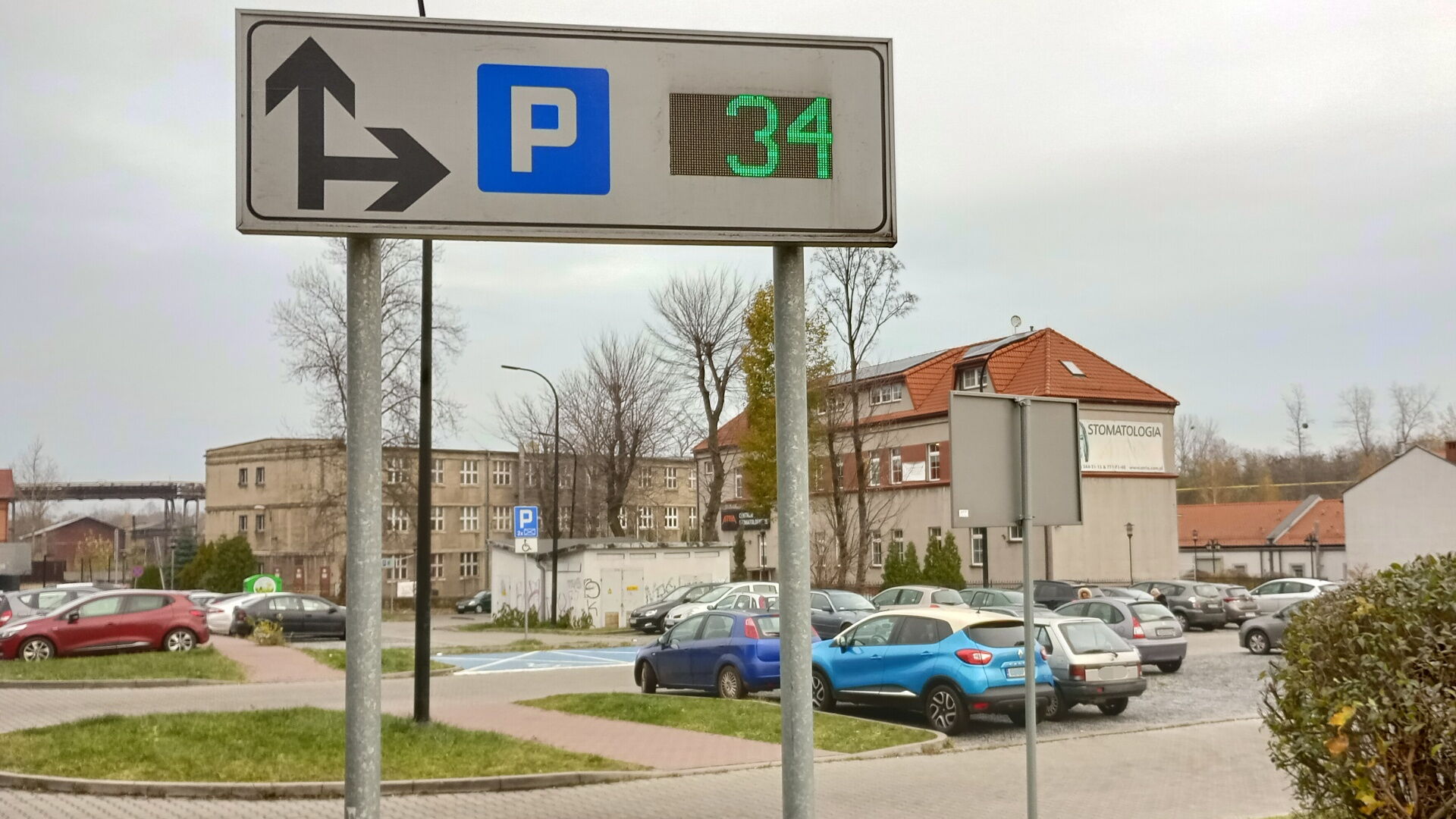 Parking z tablicą wyświetlającą liczbę wolnych miejsc. W tle budynki.