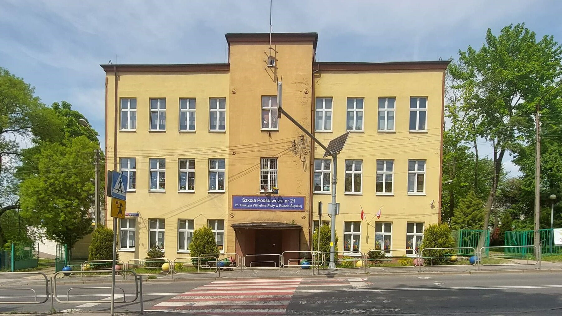 Jezdnia, przejście dla pieszych, budynek z napisem "Szkoła Podstawowa nr 21 w Rudzie Śląskiej", drzewa.