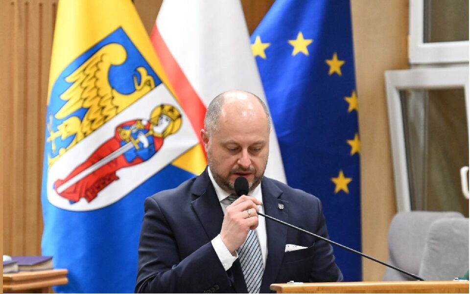 w pomieszczeniu przy mównicy mężczyzna przemawia do mikrofonu, w tle flaga Rudy Śląskiej i UE