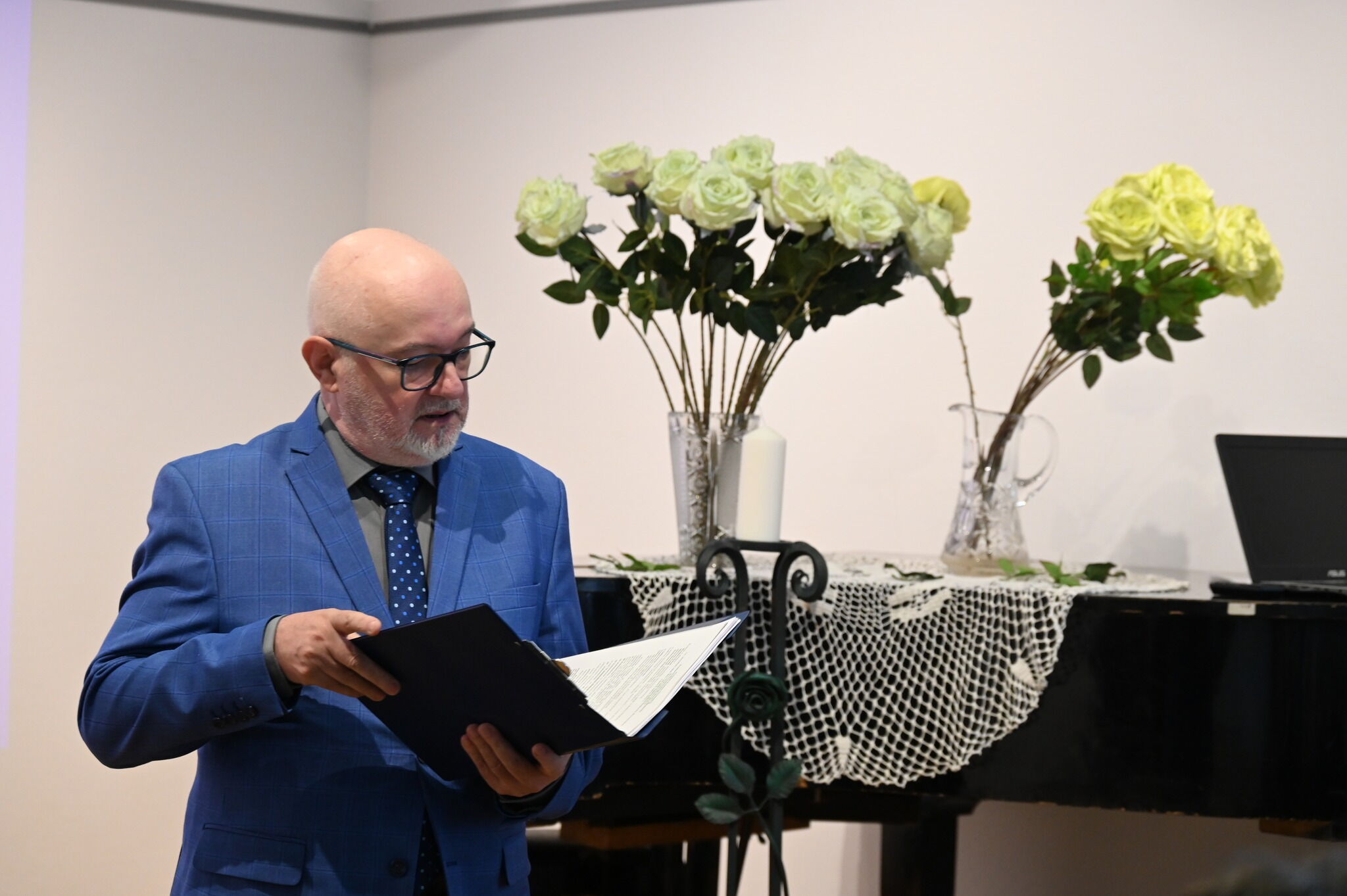 Mężczyzna z teczką przy fortepiania, na którym są ustawione zielone róze w wazonach.