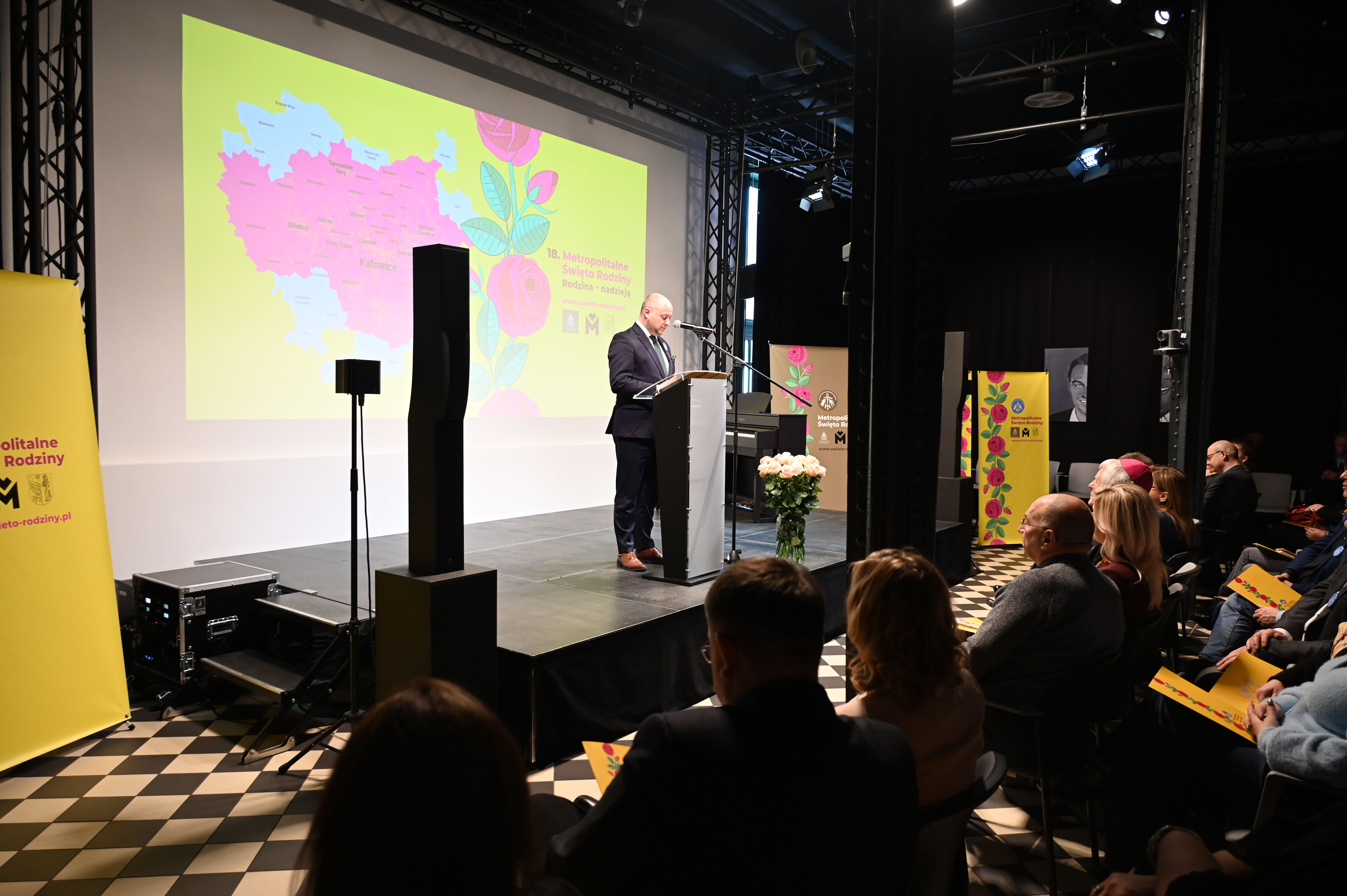 w pomieszczeniu mężczyzna stoi na scenie  przy mównicy i przemawia do mikrofonu. Na przeciw widownia