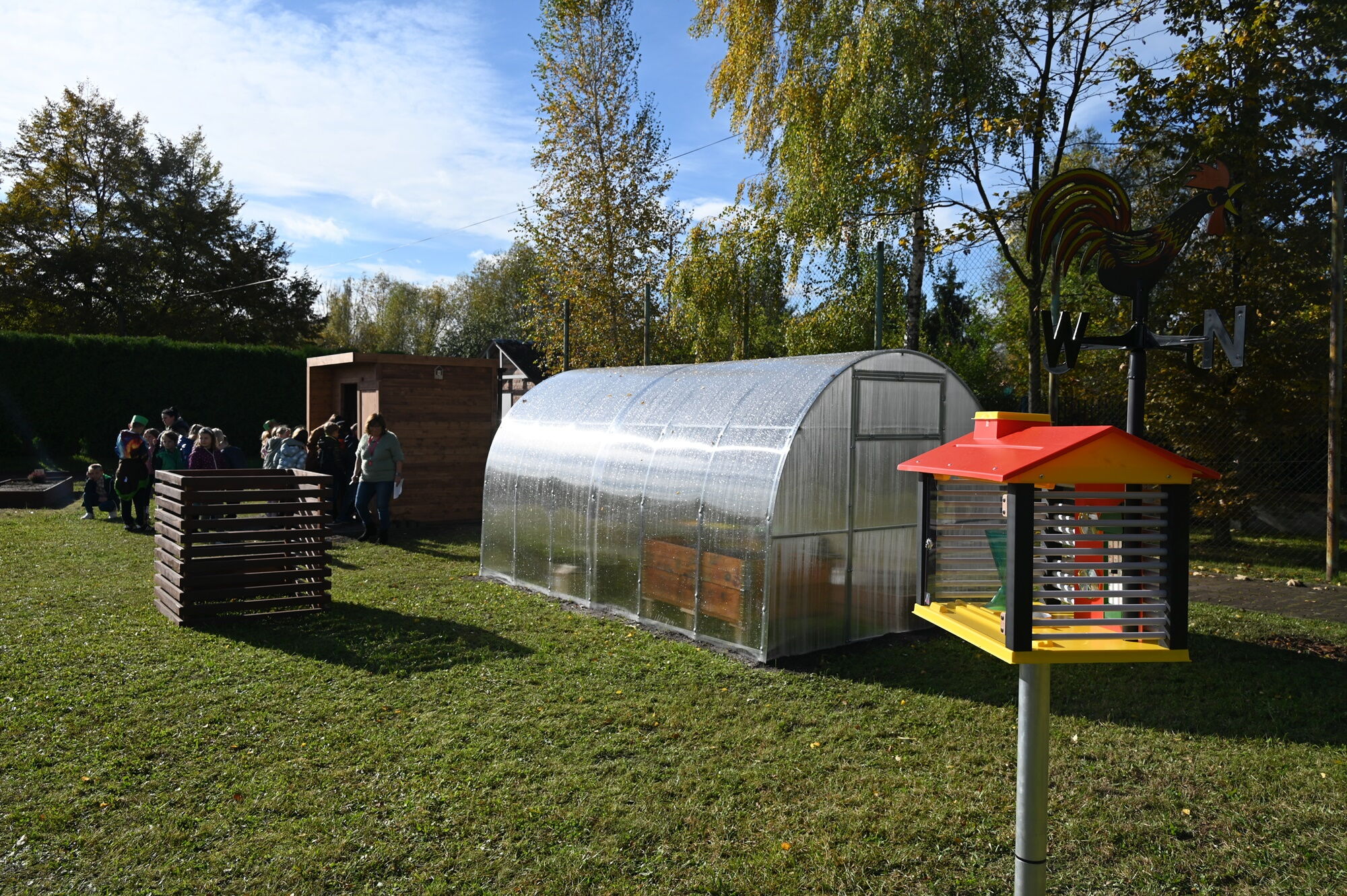 Ogródek. Na pierwszym planie kolorowy domek. W tle szklarnia, drewniany kompostownik oraz drewniany domek. Z tyłu stoi grupa dzieci.