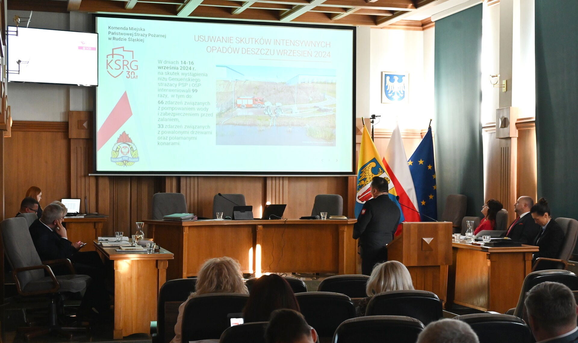 Sala sesyjna - na ekranie prezentacja ze straży pożarnej. Obok mównicy stoi mężczyzna z mikrofonem. Osoby siedzą w fotelach, widziane są od tyłu.