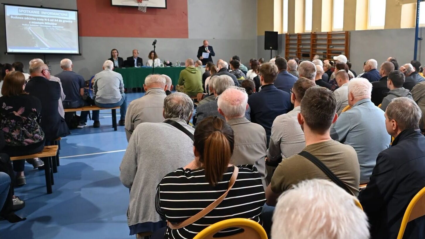 Grupa osób siedzi w sali gimnastycznej. W tle mężczyzna z mikrofonem i ekran z prezentacją.