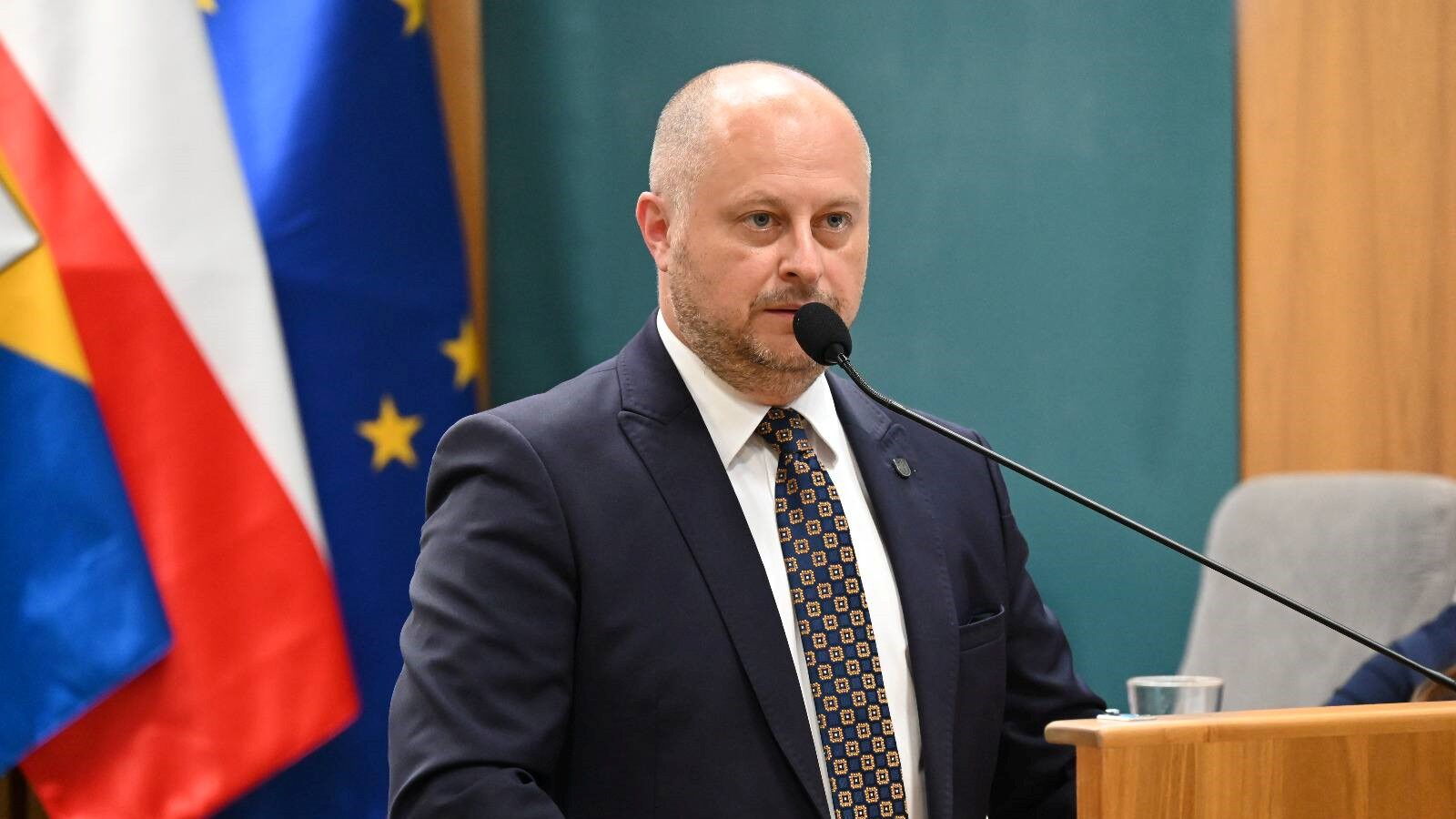 Mężczyzna w garniturze przy mównicy. W tle flagi Rudy Śląskiej, Polski i Unii Europejskiej.
