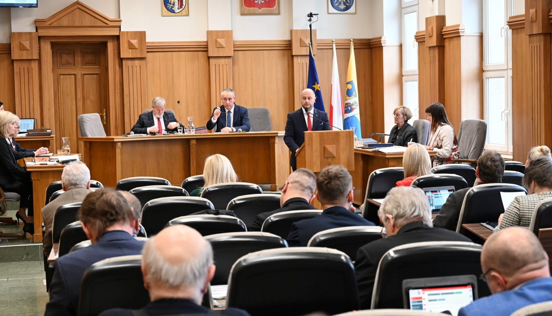 Sala sesyjna. Grupa osób siedzących na fotelach, w tle mężczyzna przy mównicy i kilka osób za stołem prezydialnym. Z tyłu flagi Unii Europejskiej, Polski, i Rudy Śląskiej.