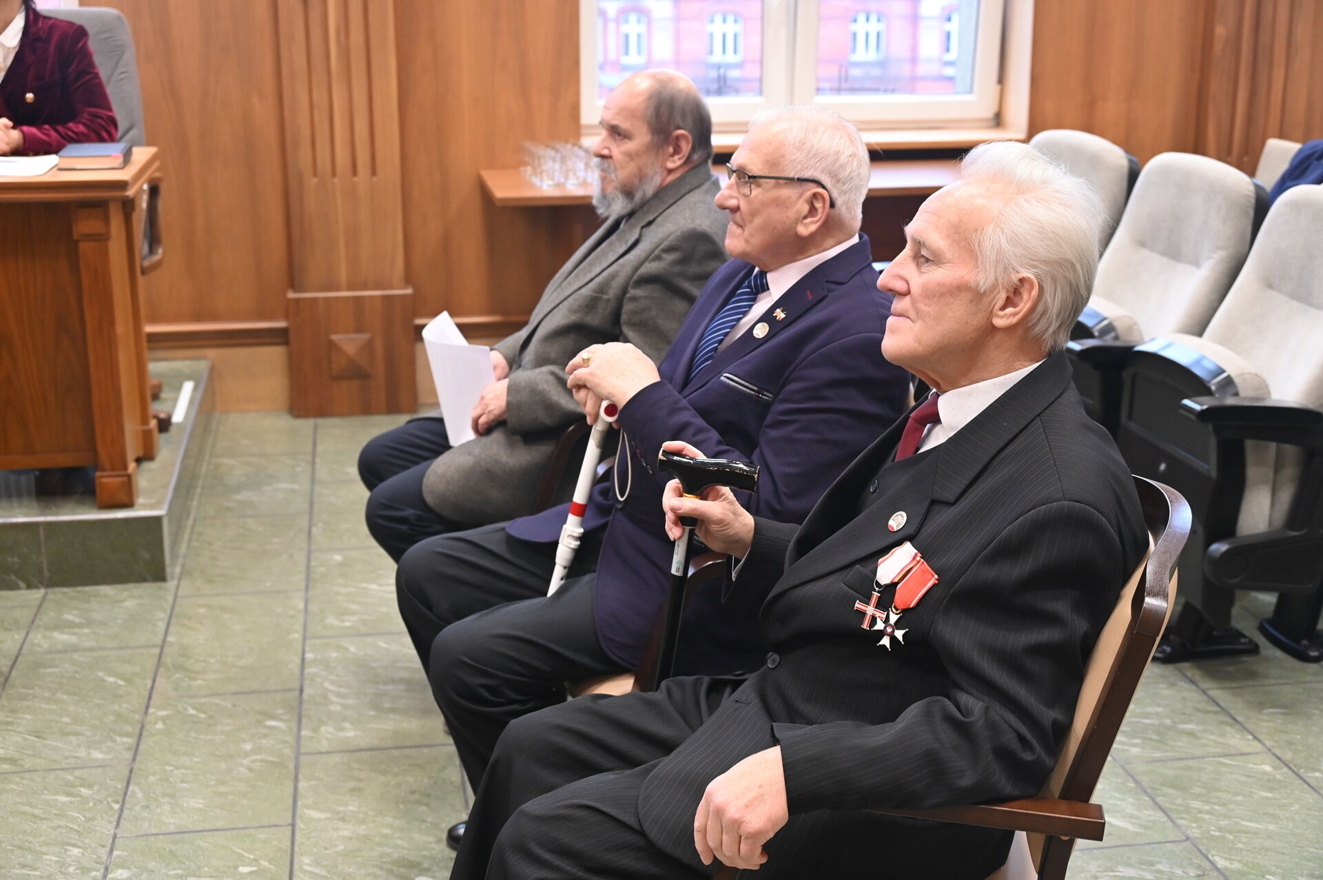 Trzech mężczyzn z garniturach siedzi na sali.