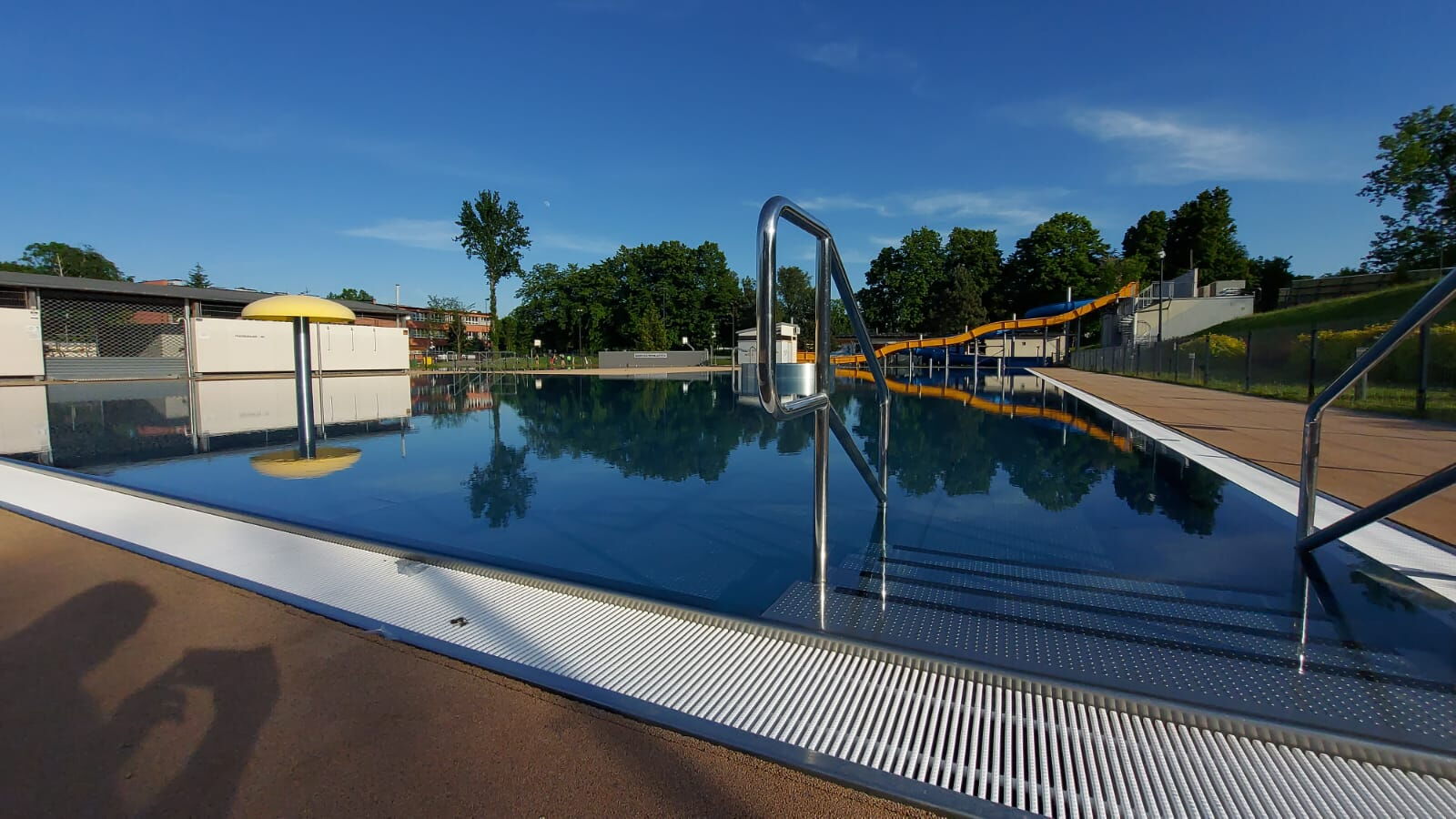 Otwarty basen letni. Na pierwszym planie barierka. W tle drzewa, budynki.