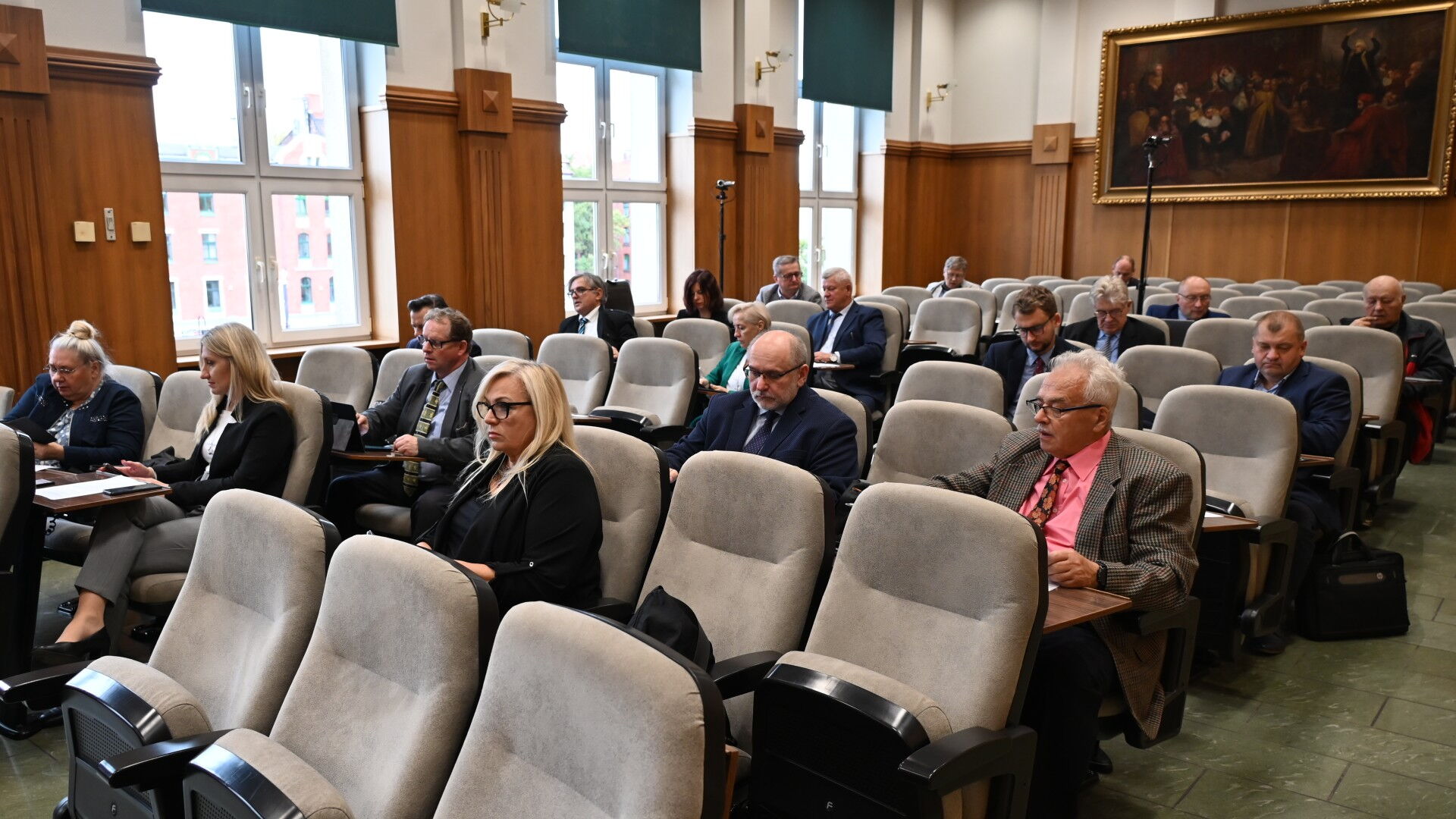 Sala obrad, grupa osób siedzącyc w fotelach, w tle obraz na ścianie.