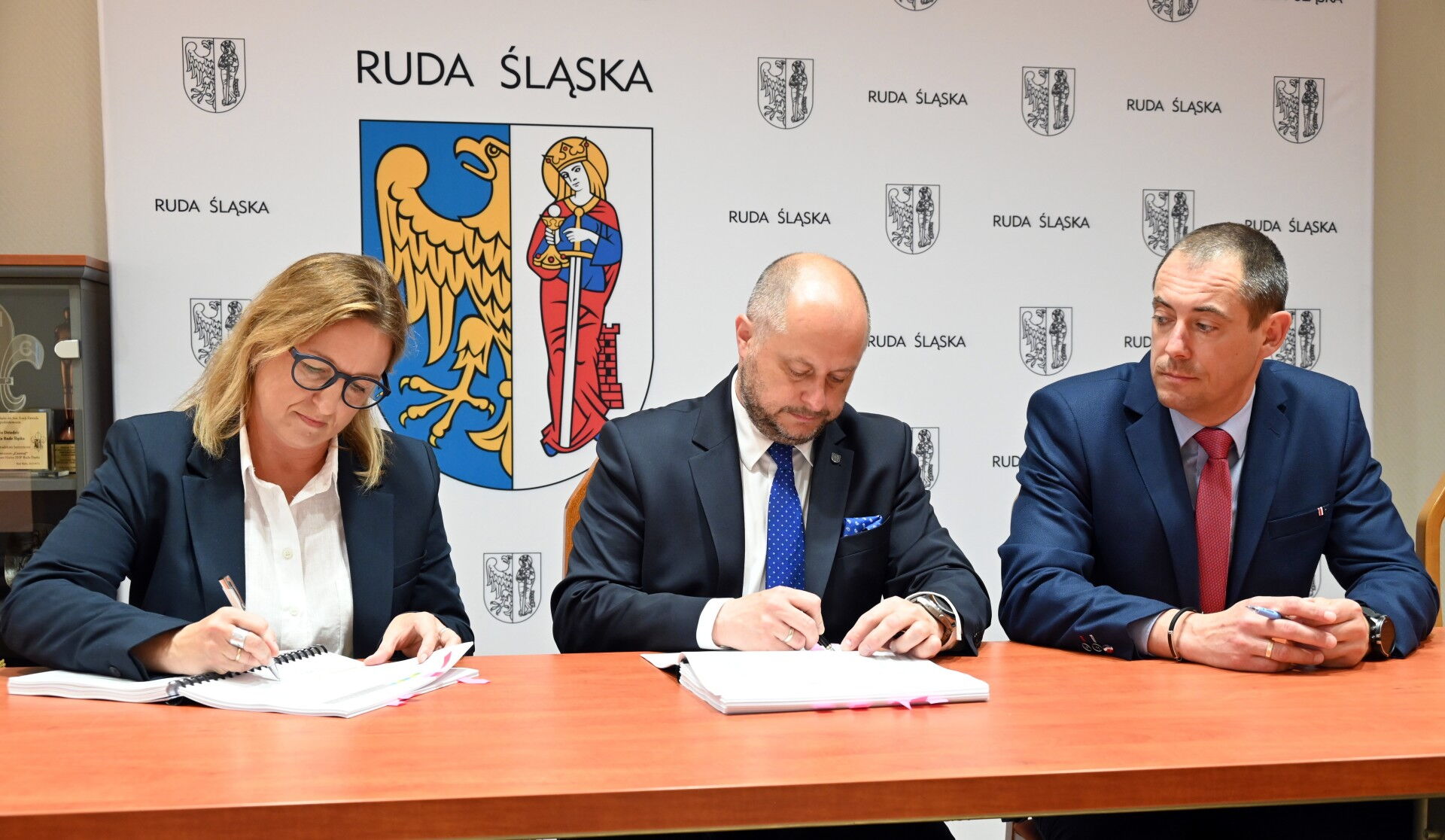 Kobieta i dwóch mężczyzn w ganiturach podpisują dokumenty za stołem, w tle ścianka z herbem Rudy Śląskiej.