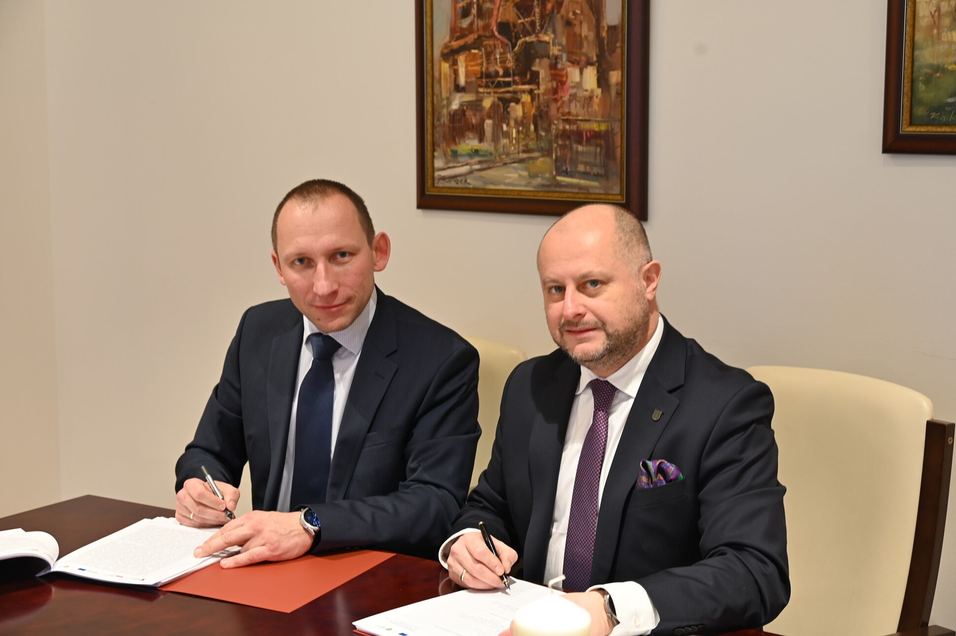 Dwaj mężczyźni w garniturach siedzą przy stole, przed nimi papiery, które podpisują.