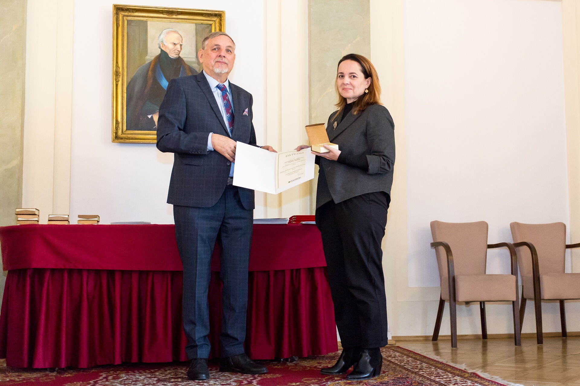 Obok siebie stoją kobieta i mężczyzna trzymając między sobą rozłożoną teczkę z dyplomem. W tle stół nakryty bordowym suknem, obraz na ścianie.