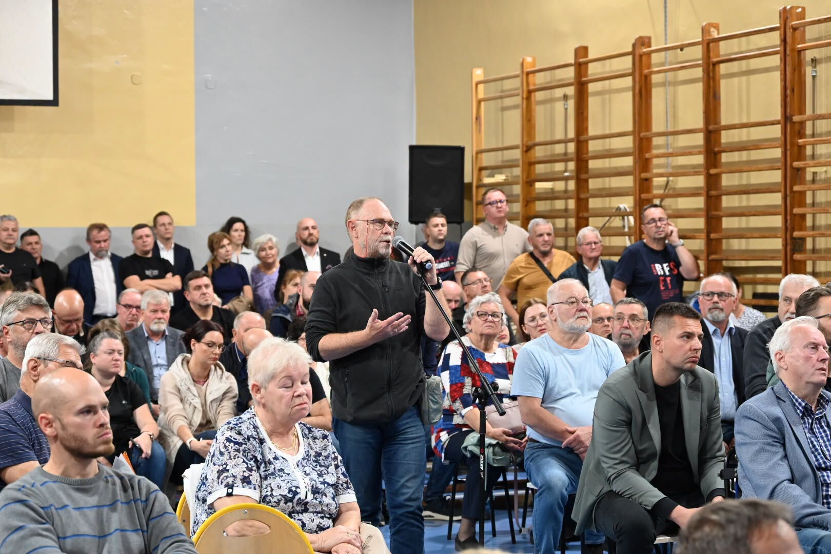 Grupa osób siedzi w sali gimnastycznej. Na środku stoi mężczyzna i mówi do mikrofonu.