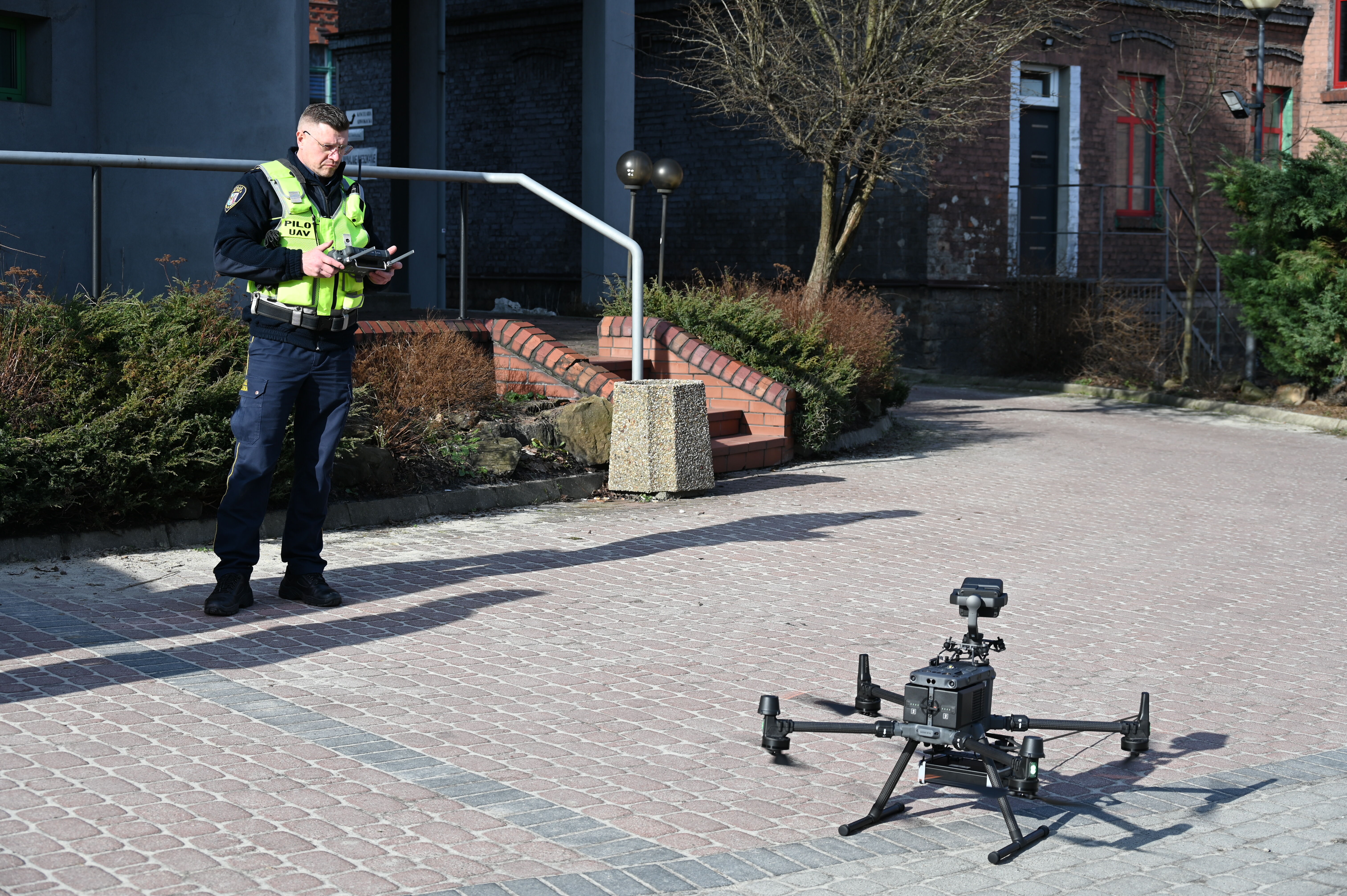 Mężczyzna w odblaskowej kamizelce steruje dronem, dron ustawiony jest na podłożu.