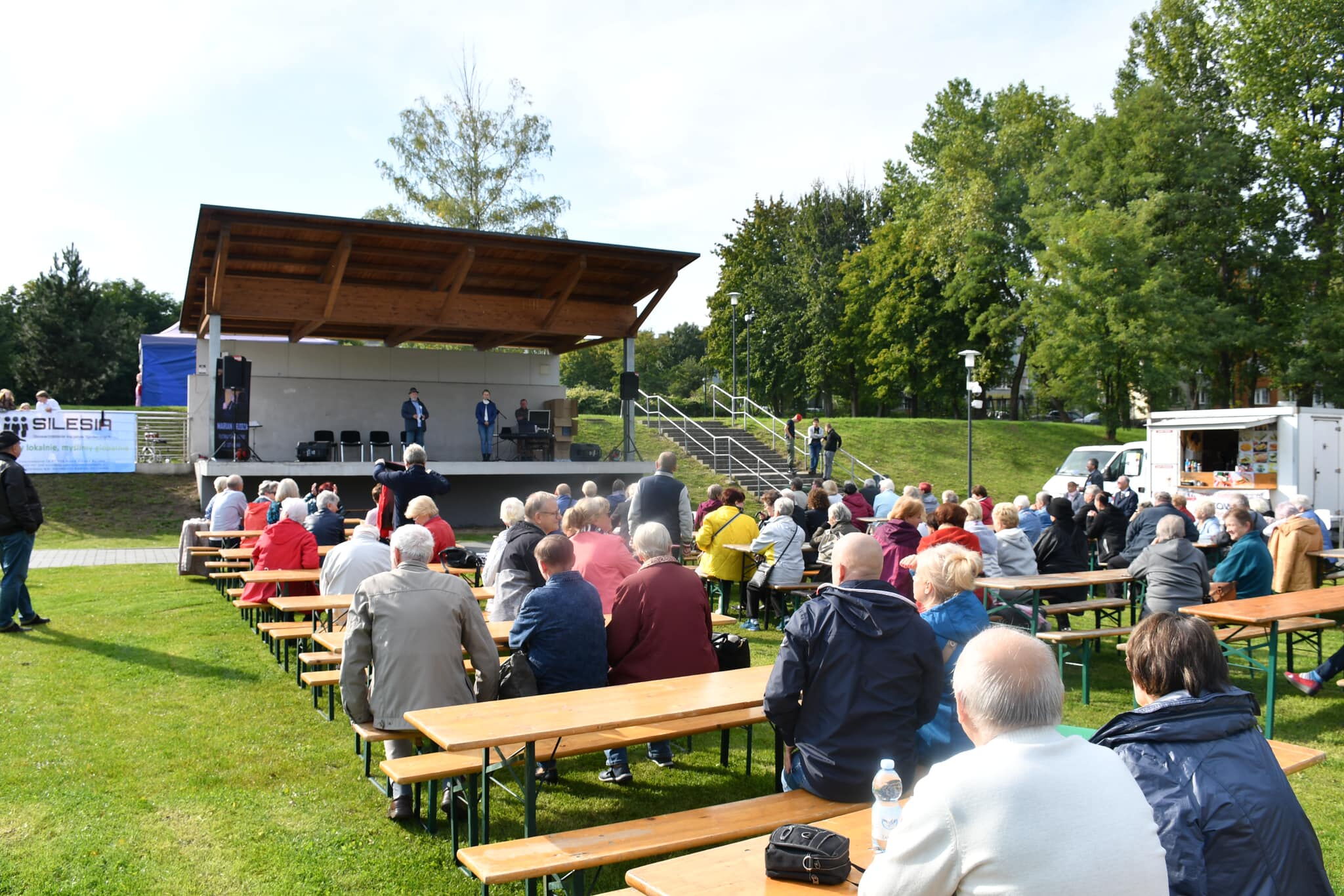 Kilkunastu seniorów siedzi na ławeczkach, w tle scena.