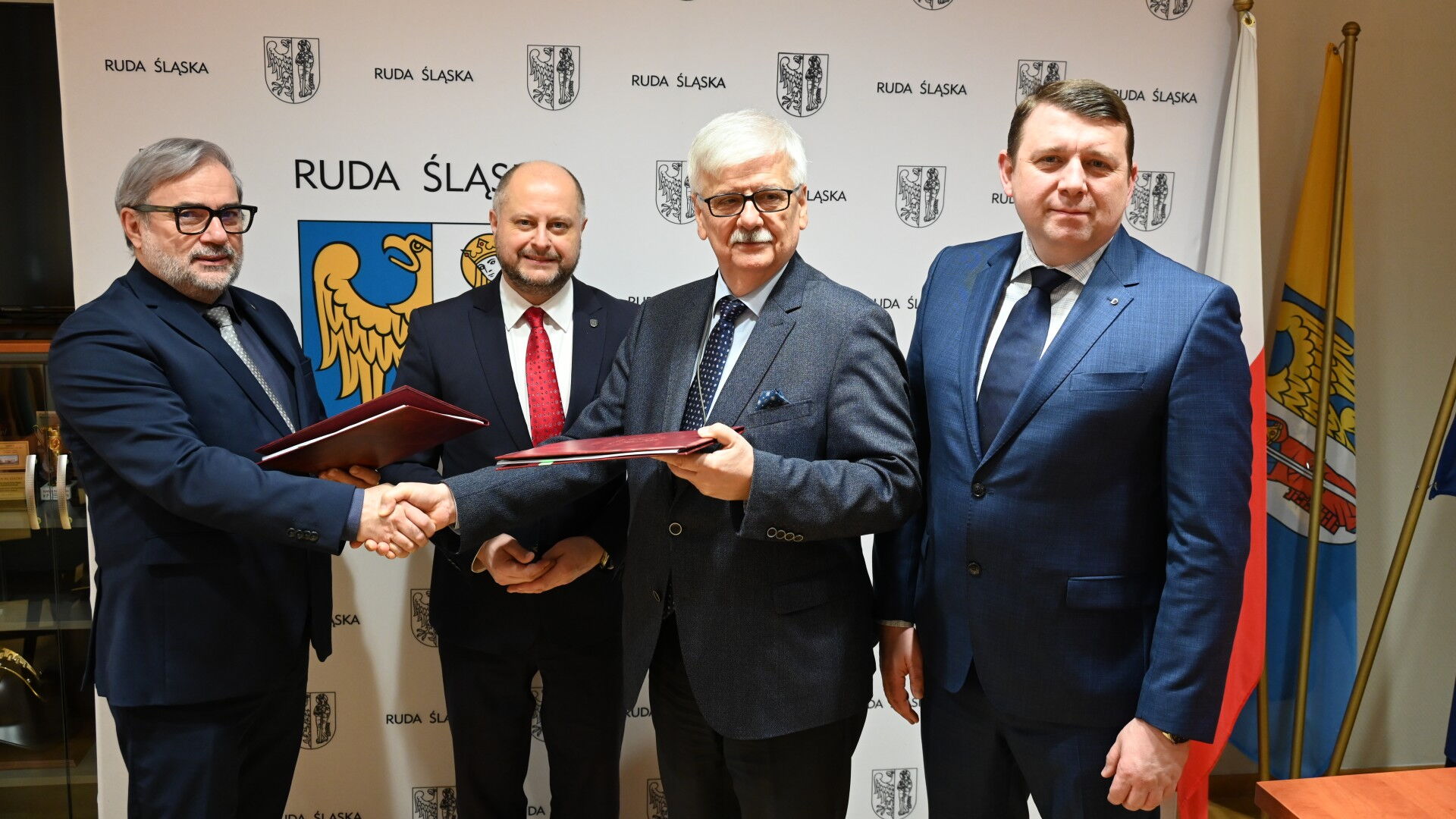 Czterech mężczyzn w garniturach. Dwóch wymienia teczki i ściska sobie dłonie. W tle ścianka z herbem Rudy Śląskiej.