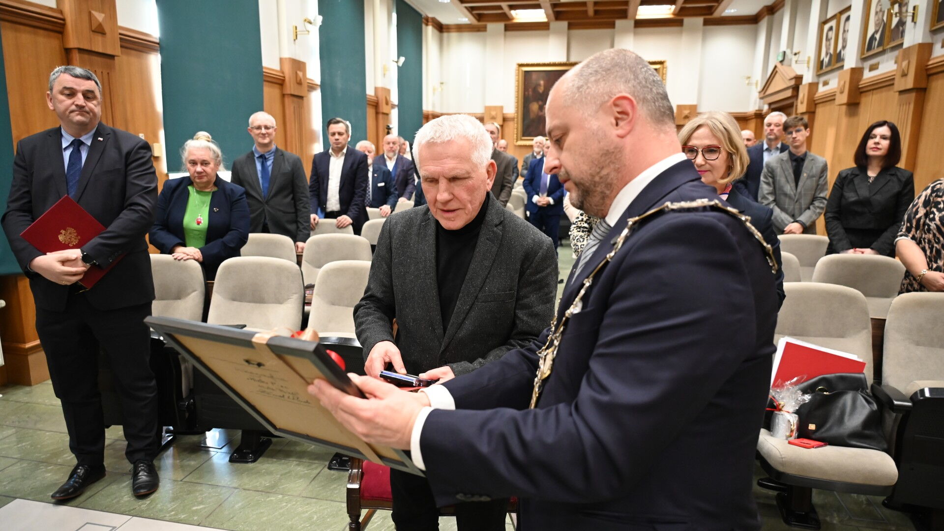 Mężczyzna wręcza obraz drugiemu mężczyźnie. W tle grupa osób stosi w sali obrad.