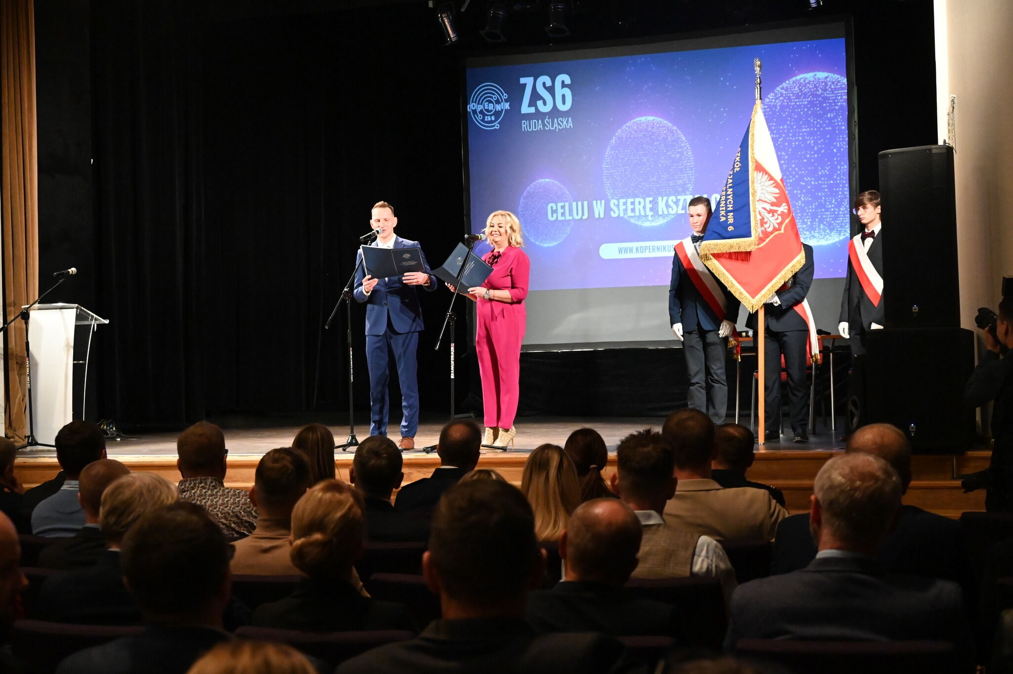 na scenie w pomieszczeniu stoja dwie osoby mówiące do mikrofonu przed publicznością, z prawej strony poczet sztandarowyny