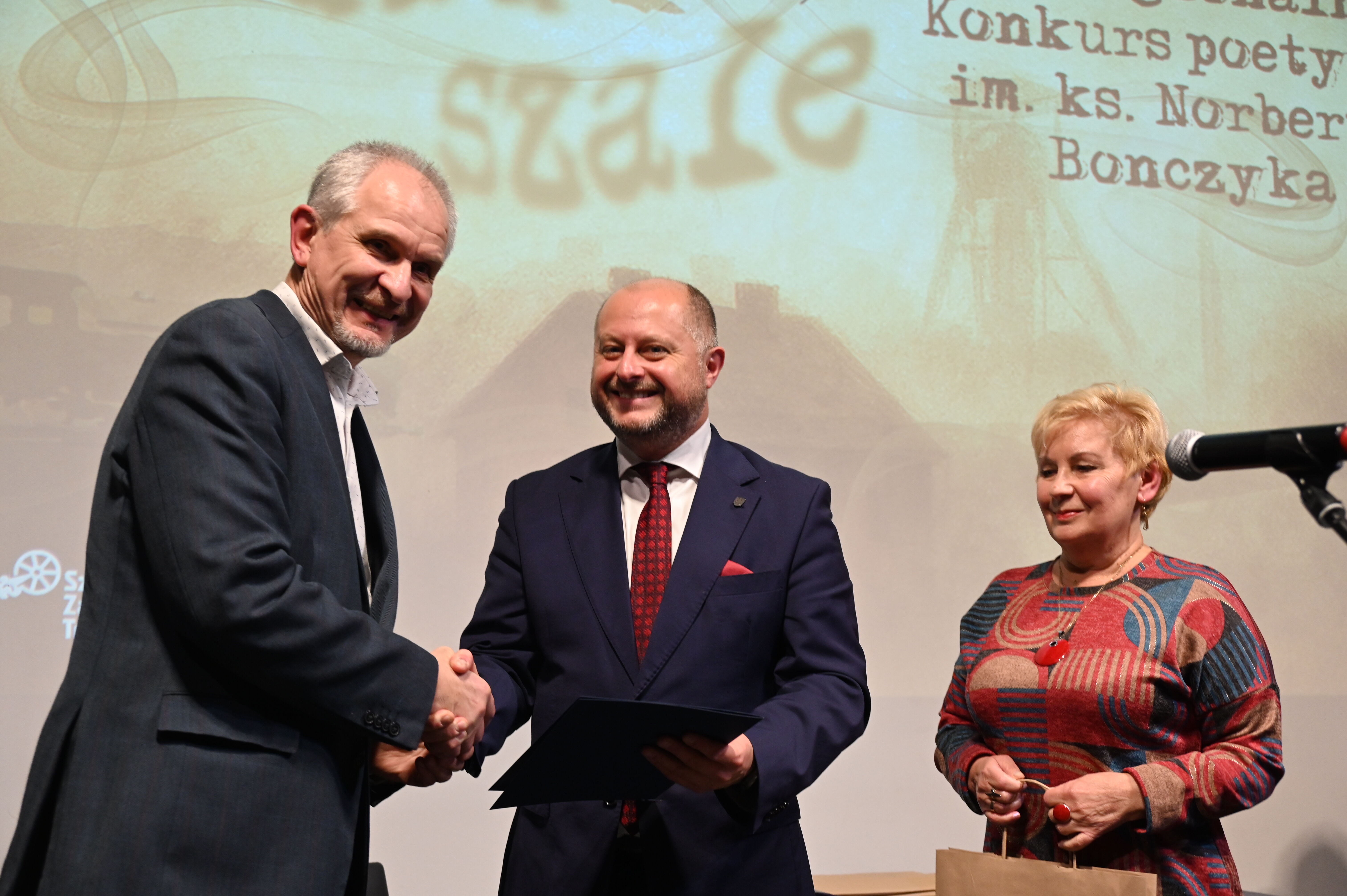 na zdjęciu trzy osoby, dwóch mężczyzn podaje sobie dłonie, obok kobieta