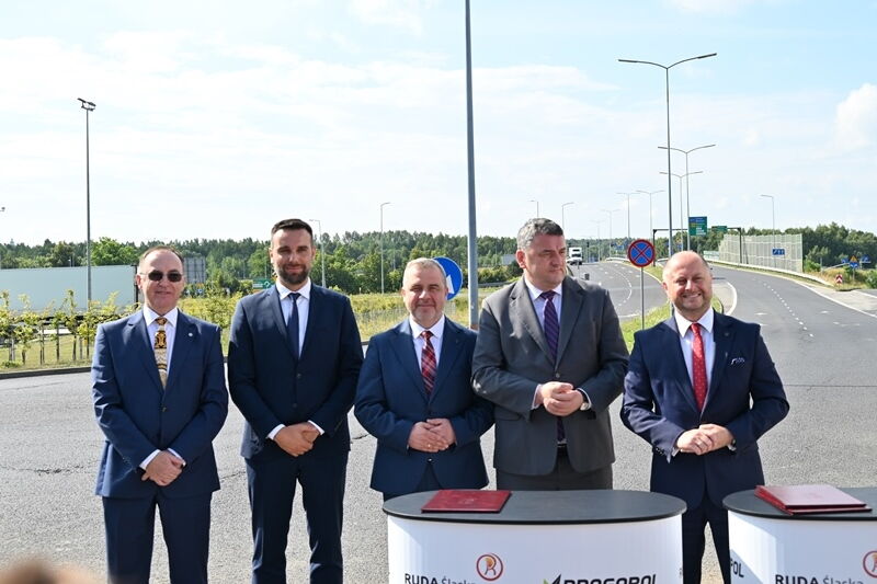 Pięciu mężczyzn w garniturach przy pulpitach z teczkami, w tle wielopasmowa droga.