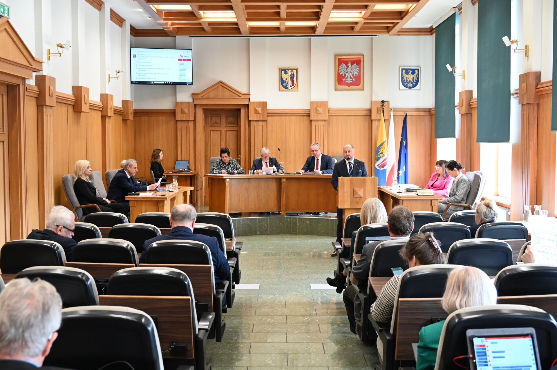 Sala sesyjna. Grupa osób siedzących na fotelach, w tle mężczyzna przy mównicy i kilka osób za stołem prezydialnym. Z tyłu flagi Unii Europejskiej, Polski, i Rudy Śląskiej.
