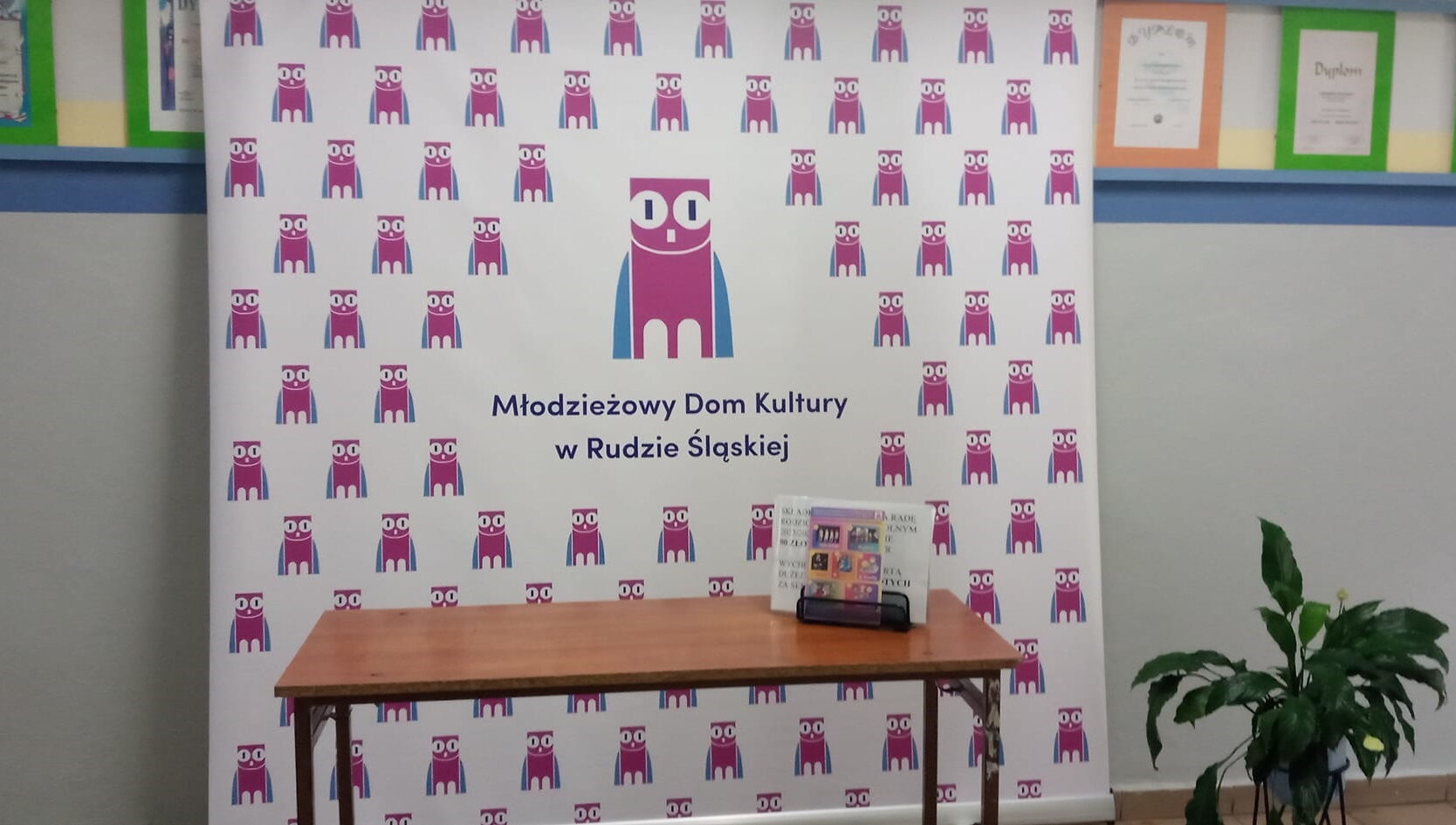 Duża plansza reklamowa z napisem Młodzieżowy dom kultury. Przed nią ławka szkolna, na niej stoi ekspozytor z ulotkami. Po prawej stronie kwiat doniczkowy.