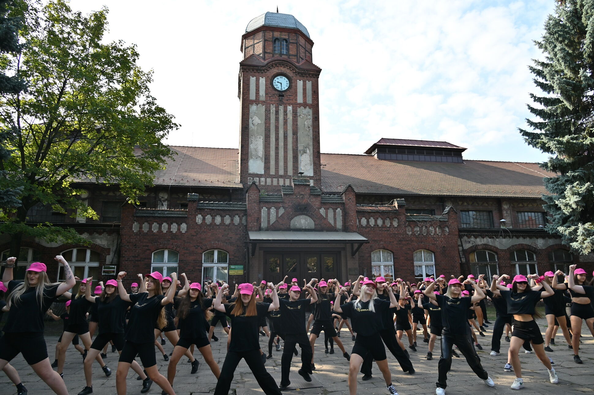 Duża grupa młodych ludzi ubranych na czarno z różowymi czapeczkami z daszkiem tańczy przed zabytkowym budynkiem z czerwonej cegły  z wieżą zegarową (KWK Bielszowice)