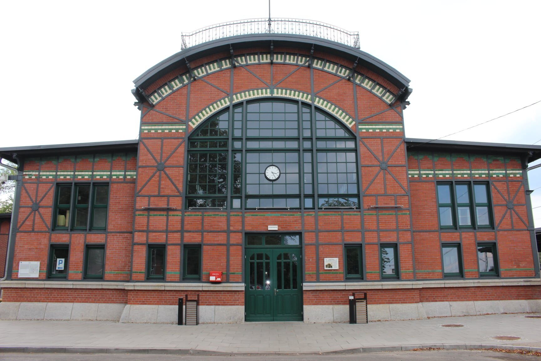 Okazały budynek dworca kolejowego z czerwonej cegły. Zielone drzwi wejściowe, centralnie powieszony zegar.