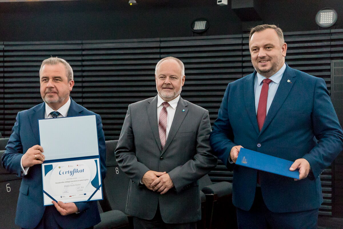 Mężczyzna w garniturze prezentuje certyfikat, obok dwóch innych mężczyzn w garniturach.