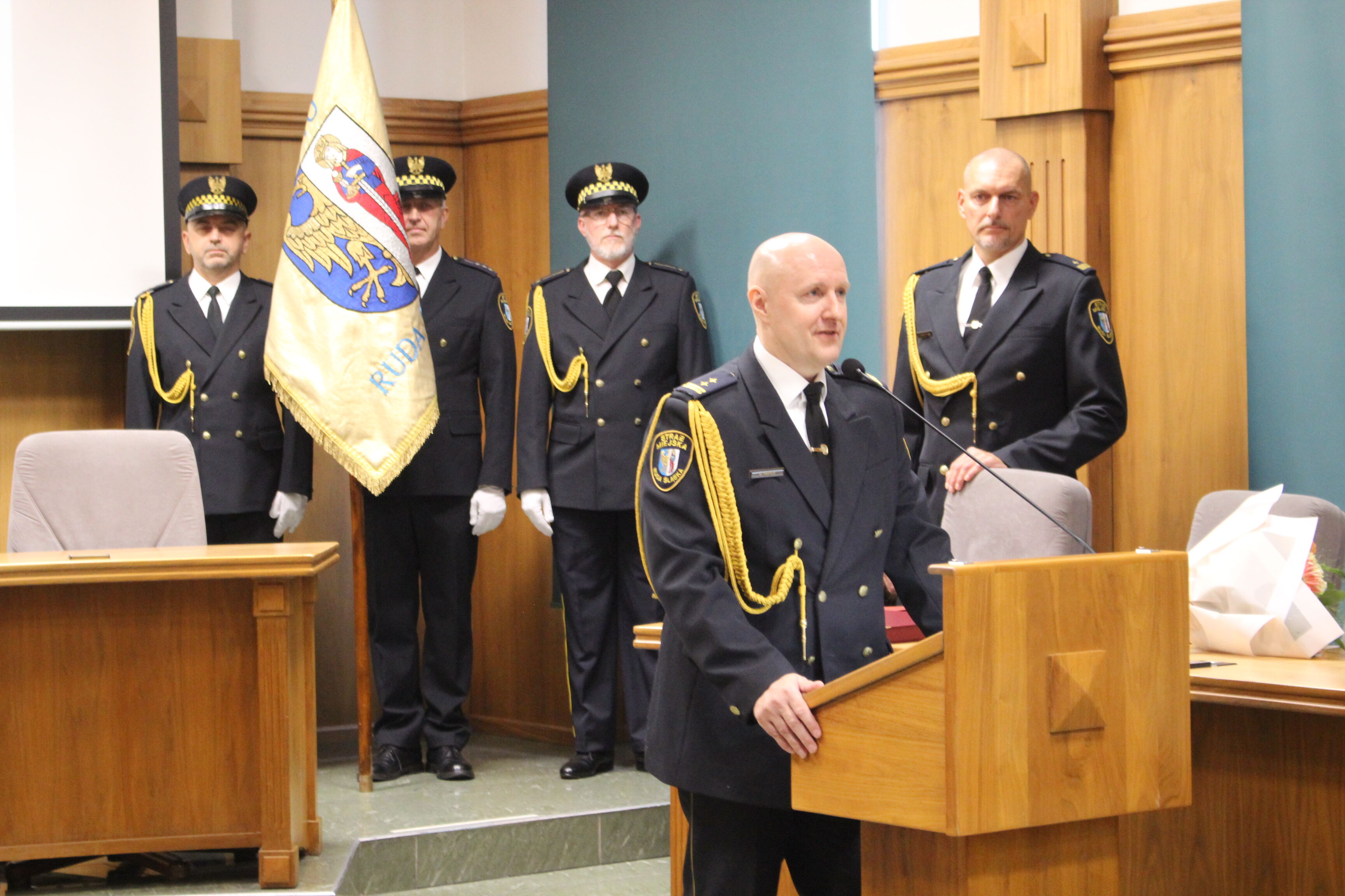 Strażnik miejski przy mównicy, za nim kilku strażników i sztandar straży miejskiej