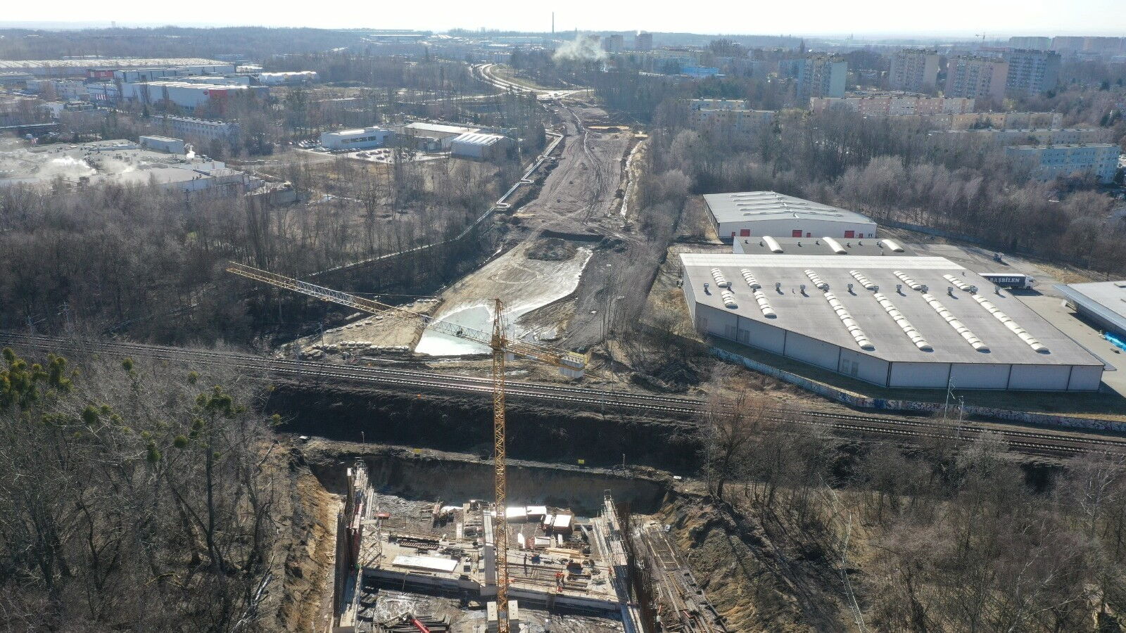 Widok z lotu ptaka: droga w budowie, hale magazynowe, budynki mieszkalne, drzewa.