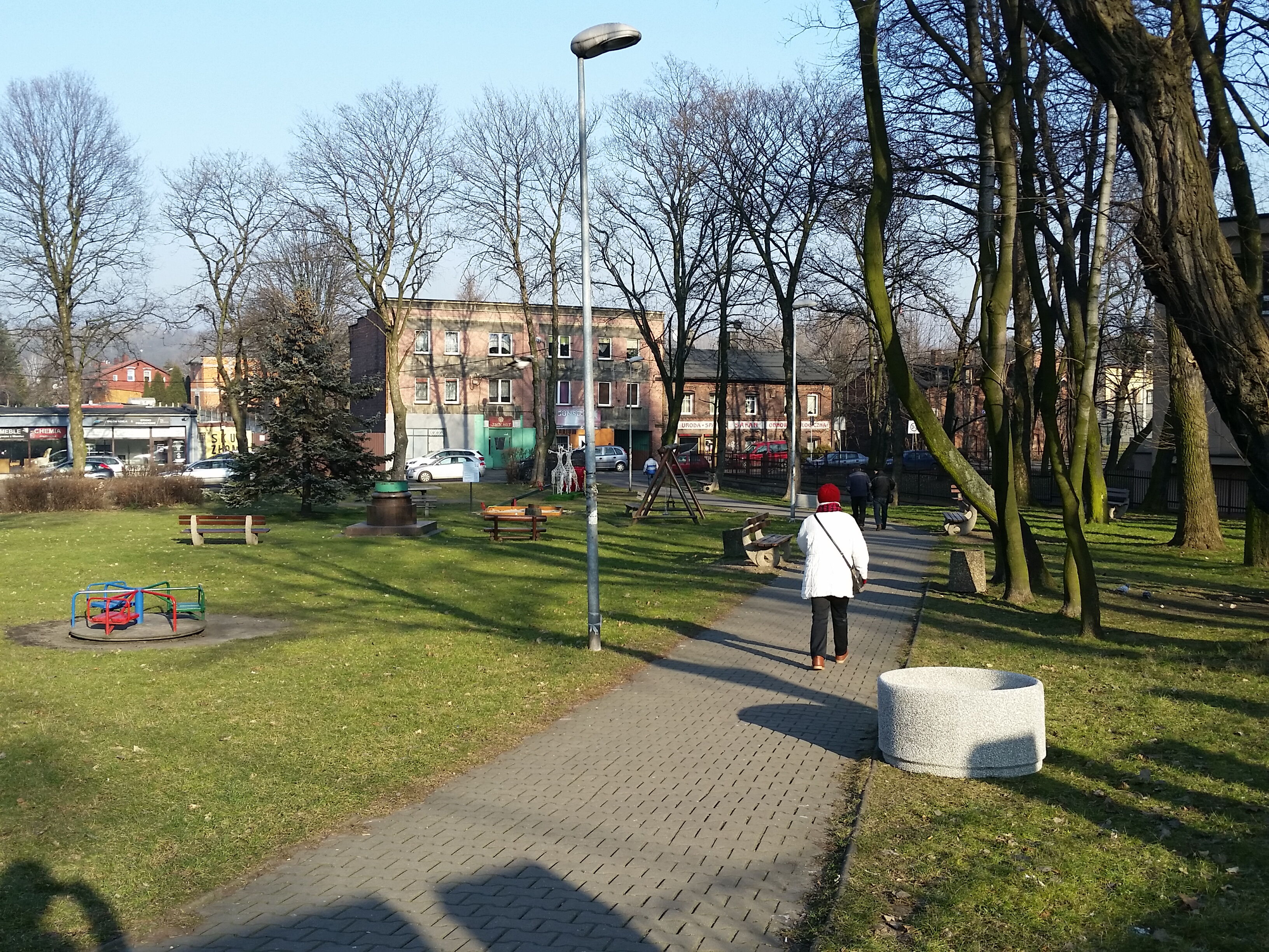 zielony skwer z ławkami i huśtwkami, przechodzące osoby