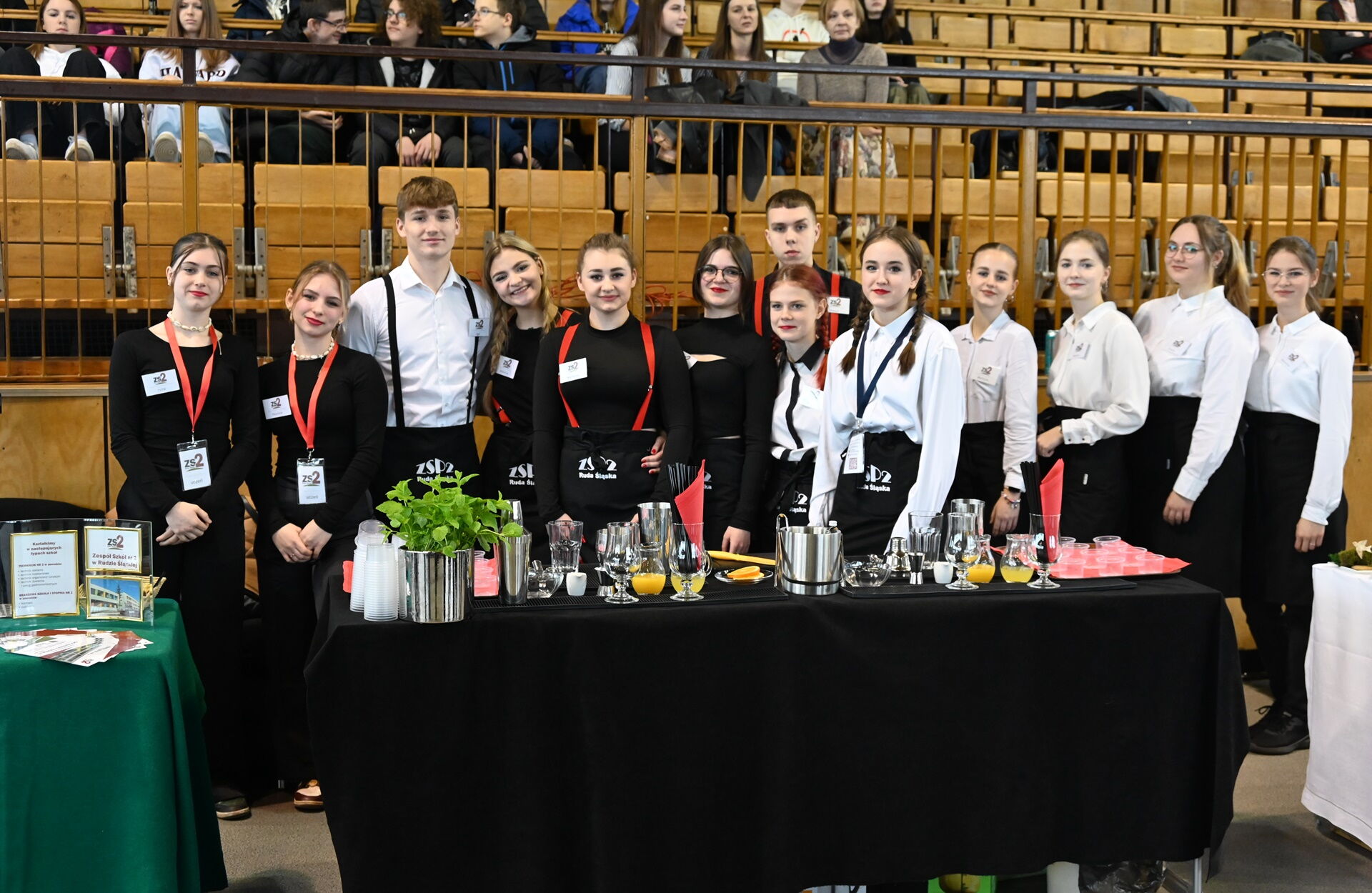 Grupa młodych ludzi - dziewczyn i chłopców stoi w biało-czarnych strojach za stołem.