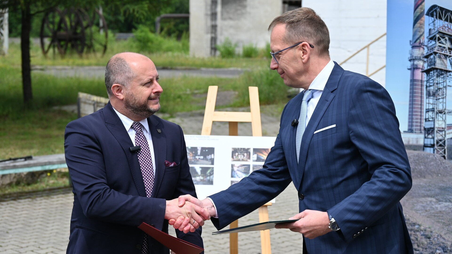 Dwaj mężczyźni w garniturach trzymają teczki i podaję sobie ręce. W tle plansza na sztaludze, zieleń i zabudowania.