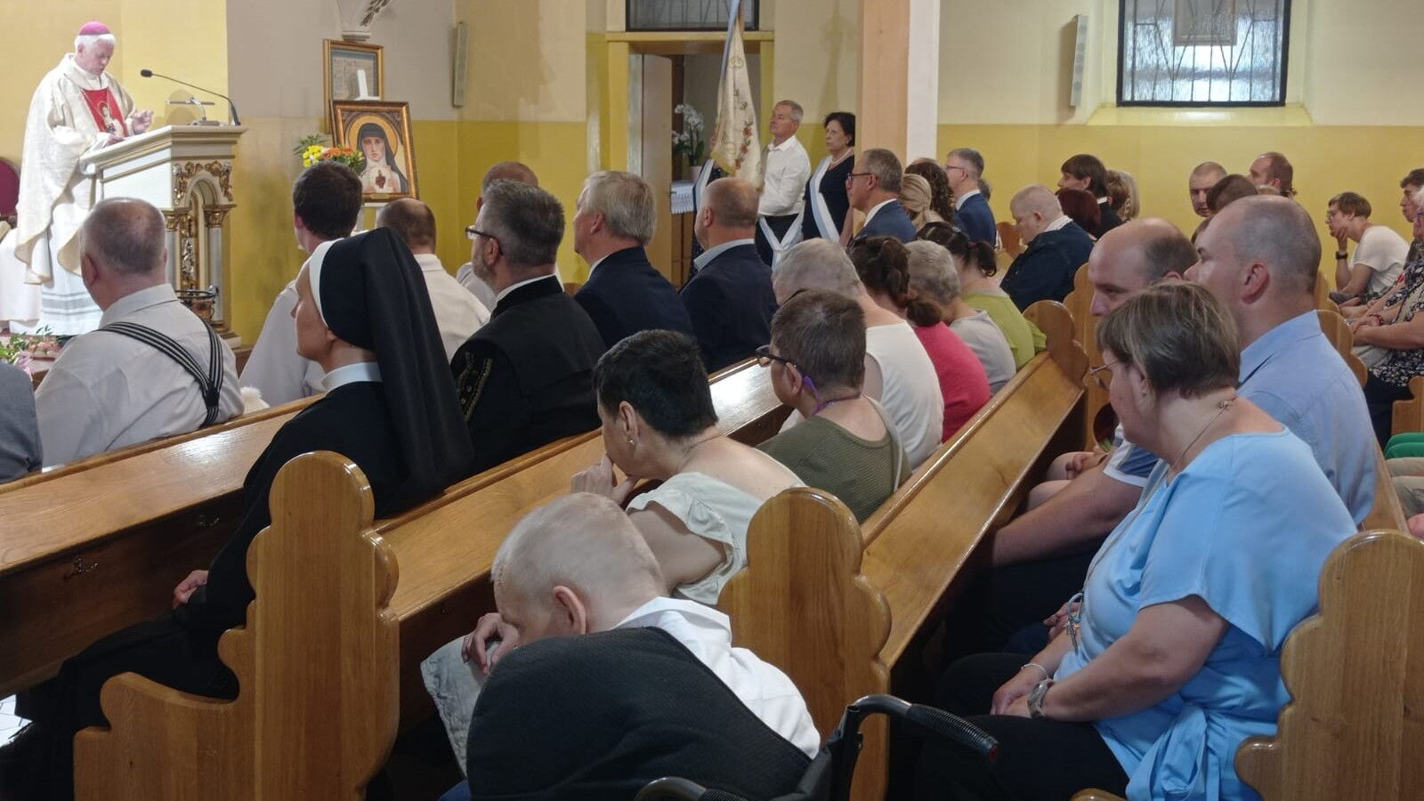 Grupa osób w kościelnych ławkach. W tle biskup przy mównicy i poczet sztandarowy.
