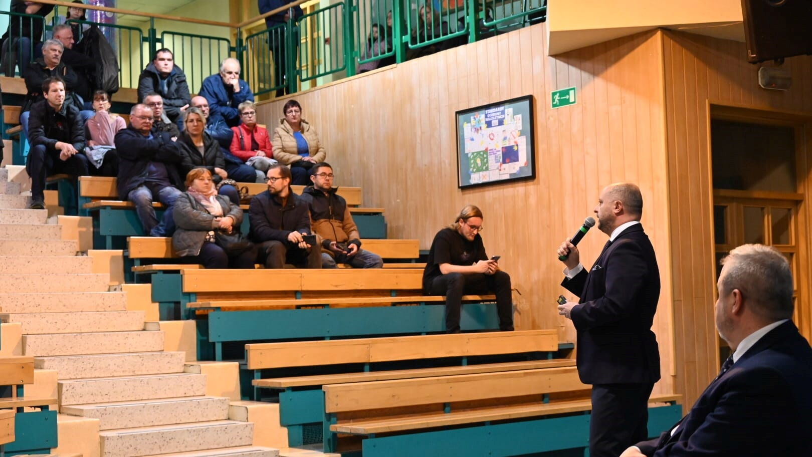 Wnętrze auli, mężczyzna z mikrofonem stoi przed grupą osób siedzących na ławkach.