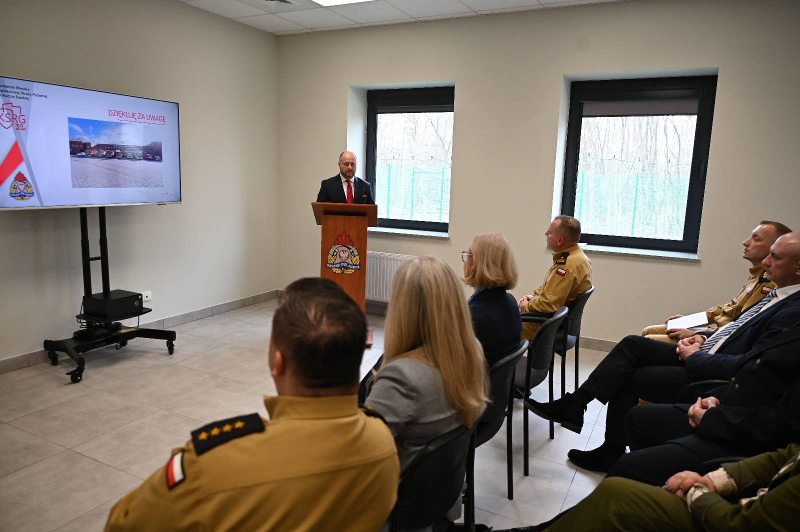 Mężczyzna stoi przy mównicy. Ludzie siedzą na krzesła.