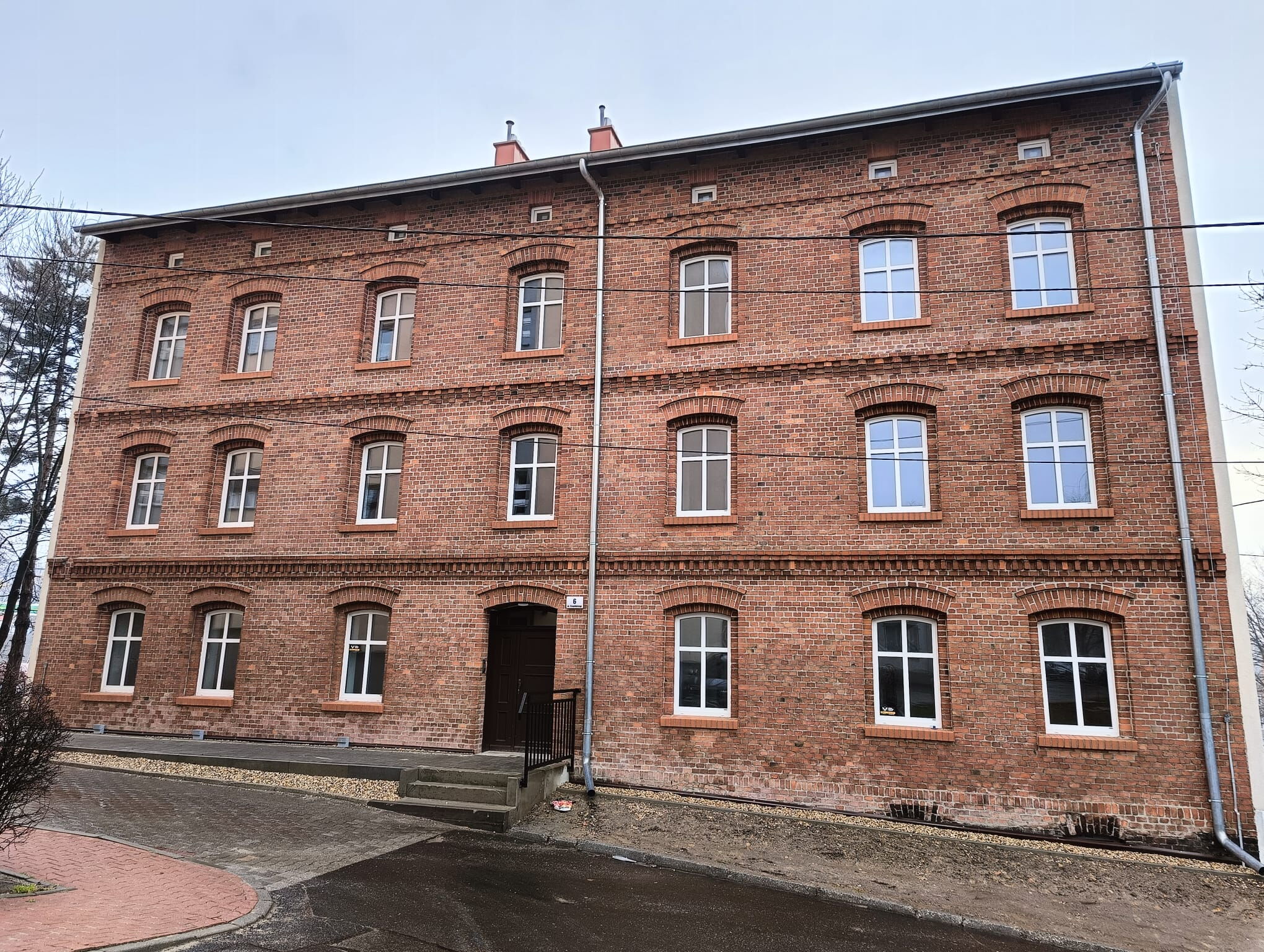 Dwupiętrowa kamienica z cegieł z białymi oknami. Wejście po kilku schodach. W tle niebo.