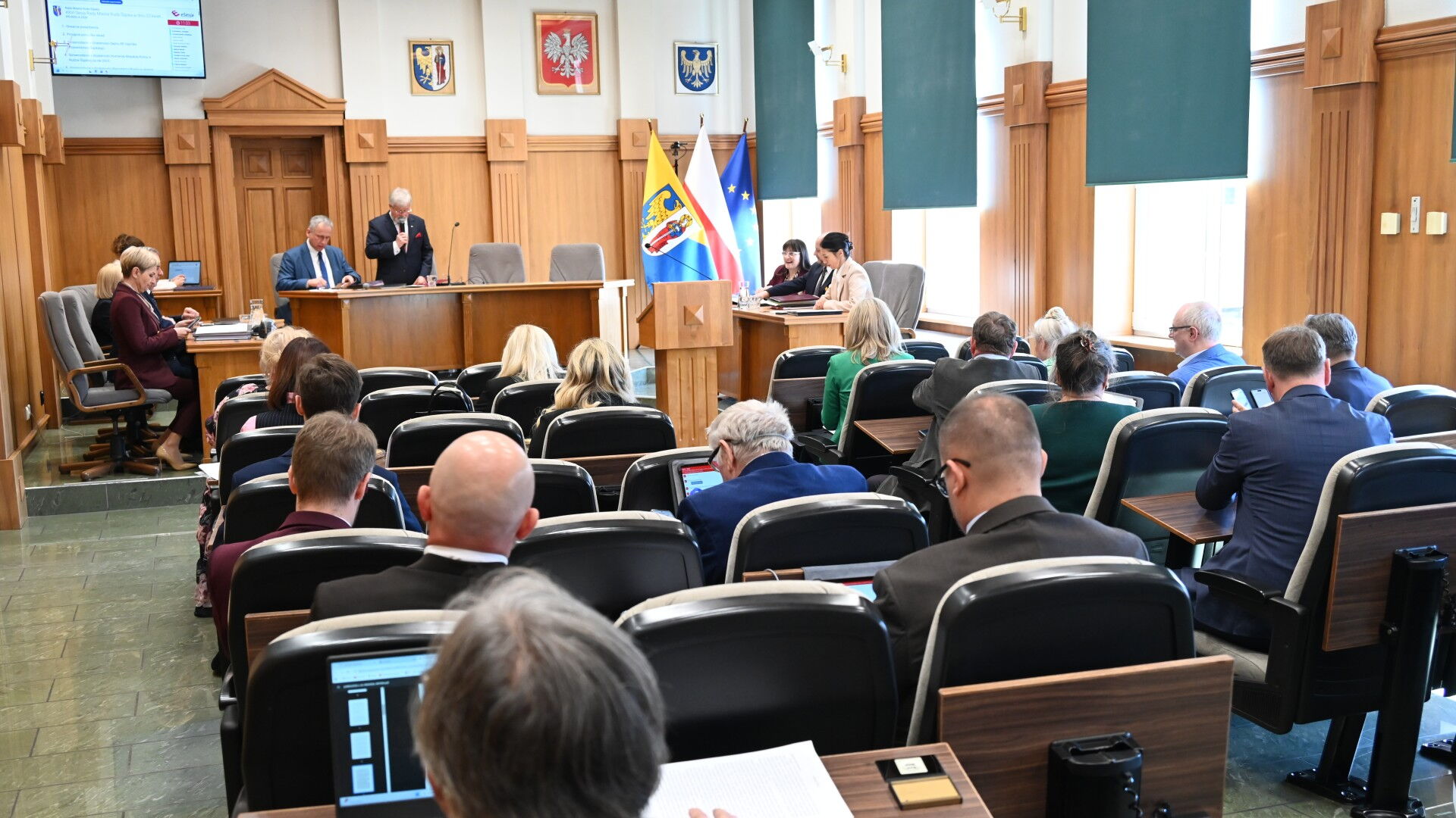 Sala sesyjna. Grupa osób siedzących na fotelach i kilka osób za stołem prezydialnym. Z tyłu flagi Unii Europejskiej, Polski i Rudy Śląskiej.