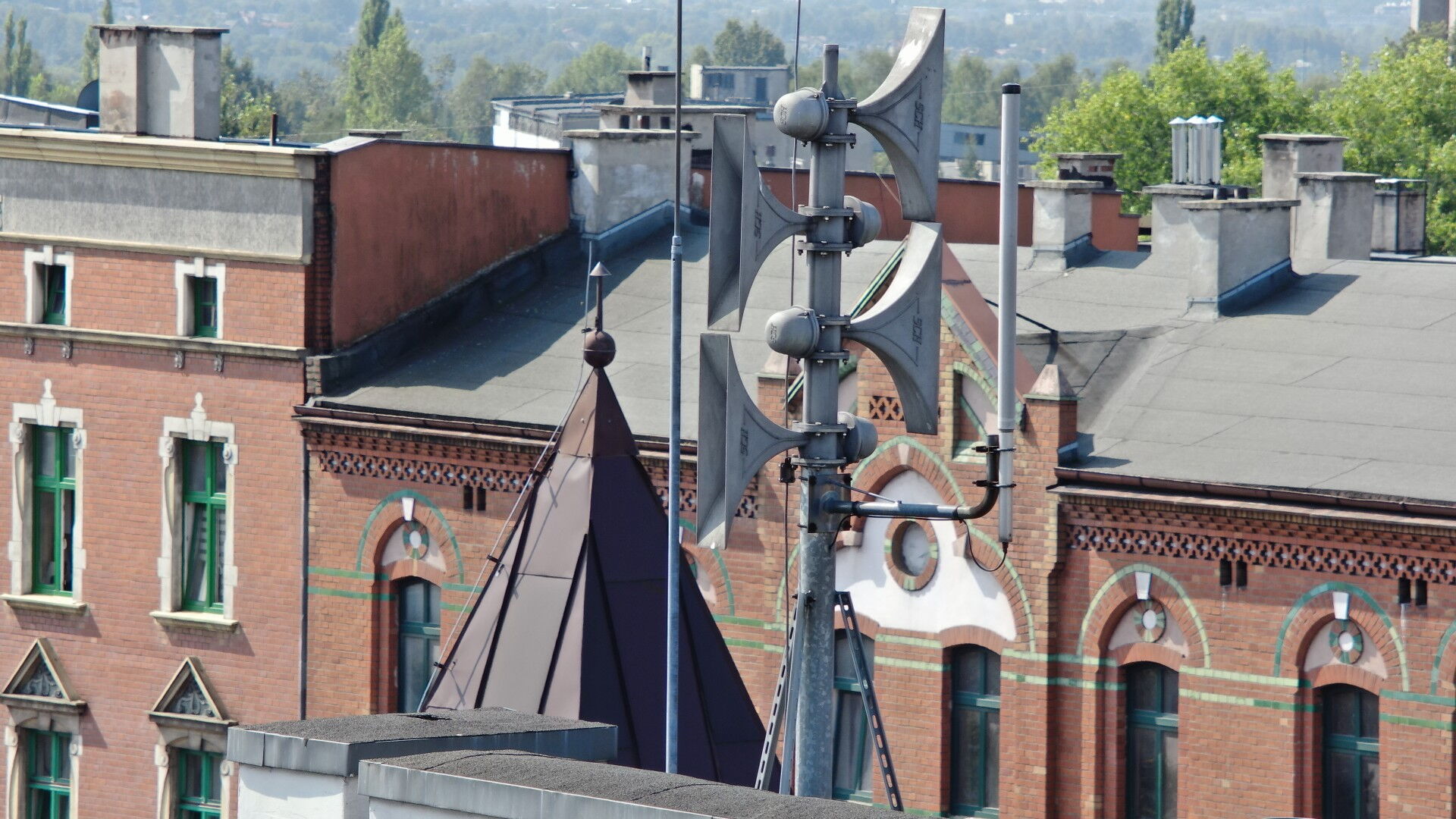 Syrena alarmowa na dachu, w tle budynki i drzewa.