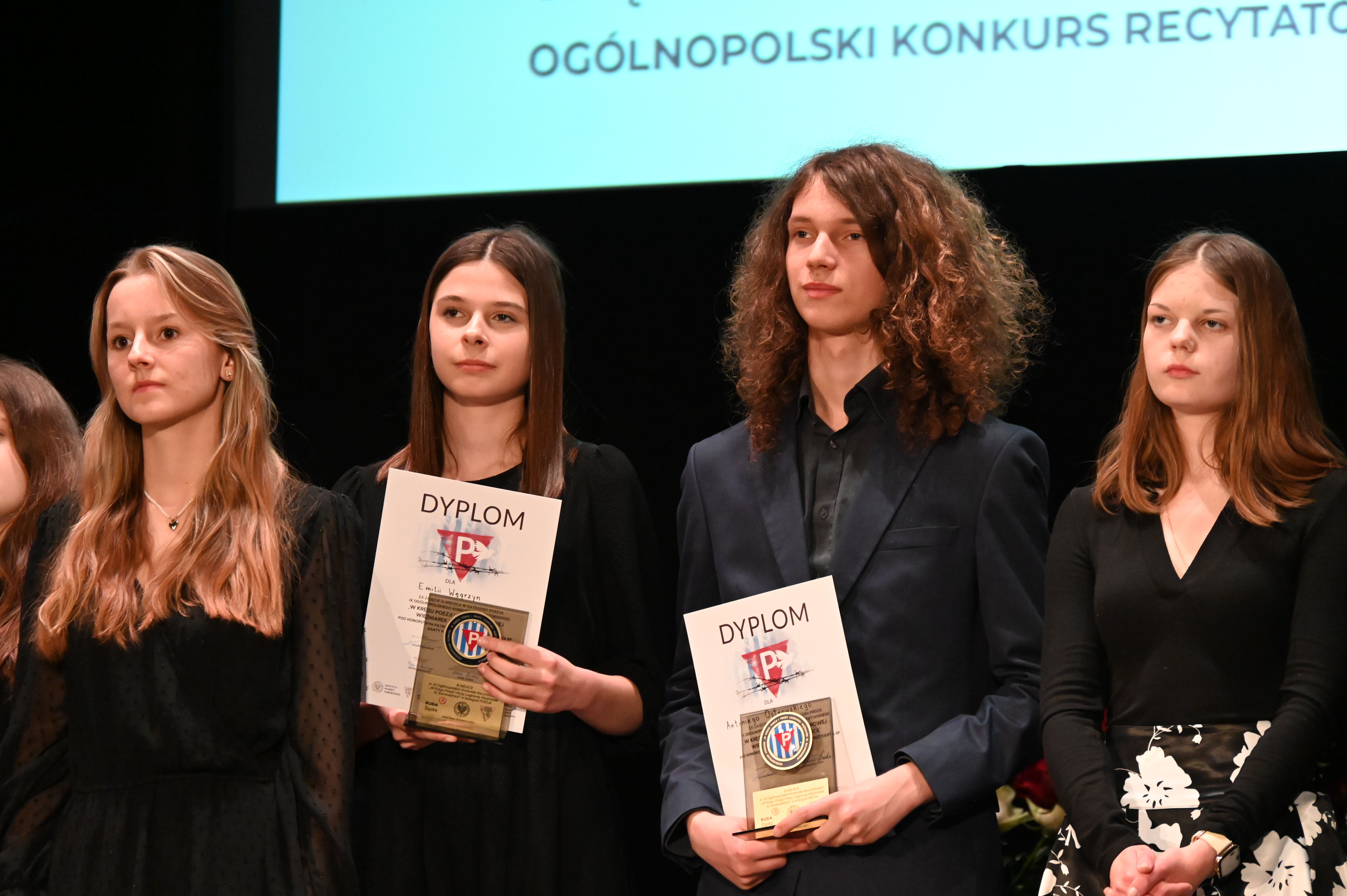 w pomieszczeniu na scenie stoją młodzi ludzie, w środku dziewczyna i chłopak trzymają dyplomy i statuetki
